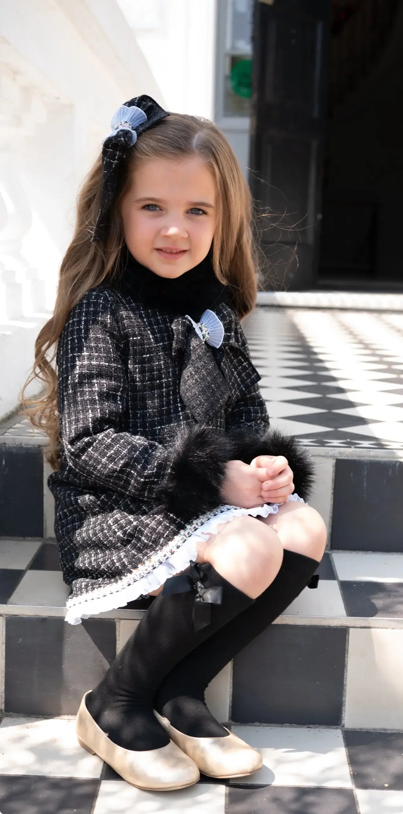 Young girl in black and white plaid dress with fur cuffs and bow sitting on black and white checkered steps