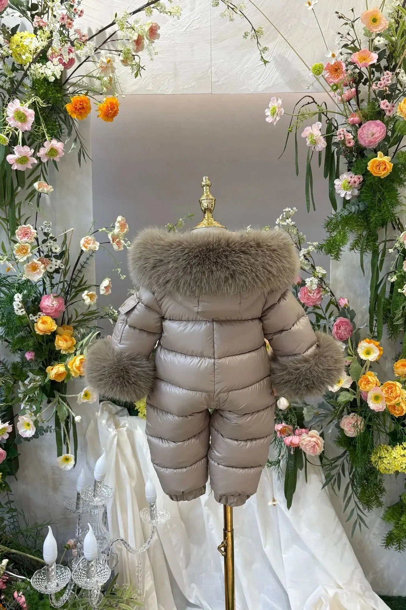 Children's beige puffer snowsuit with fur trim on collar and cuffs displayed on mannequin amid colorful floral arrangement