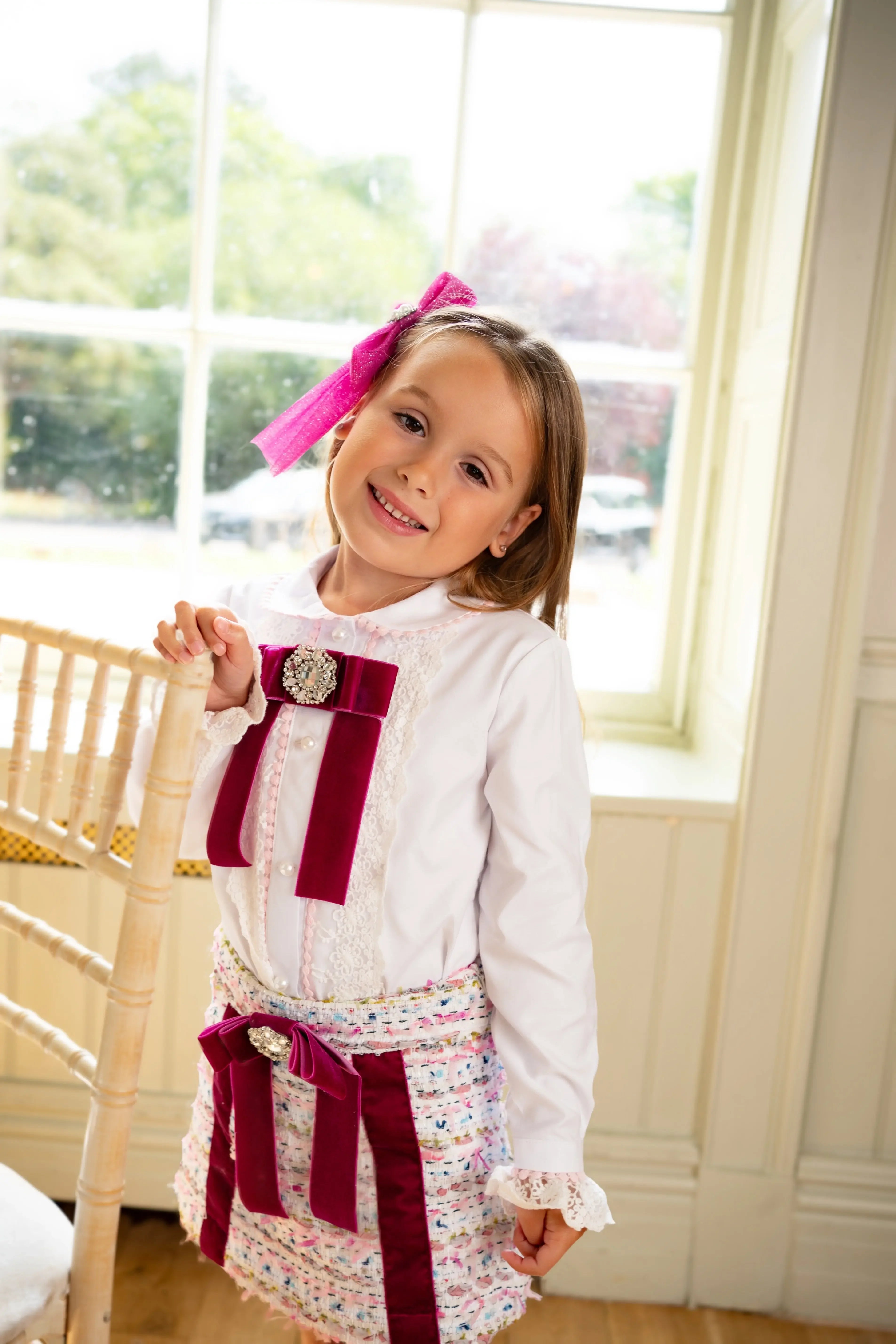 Smiling girl in white blouse and multicolor skirt with magenta bows standing by wooden chair near window