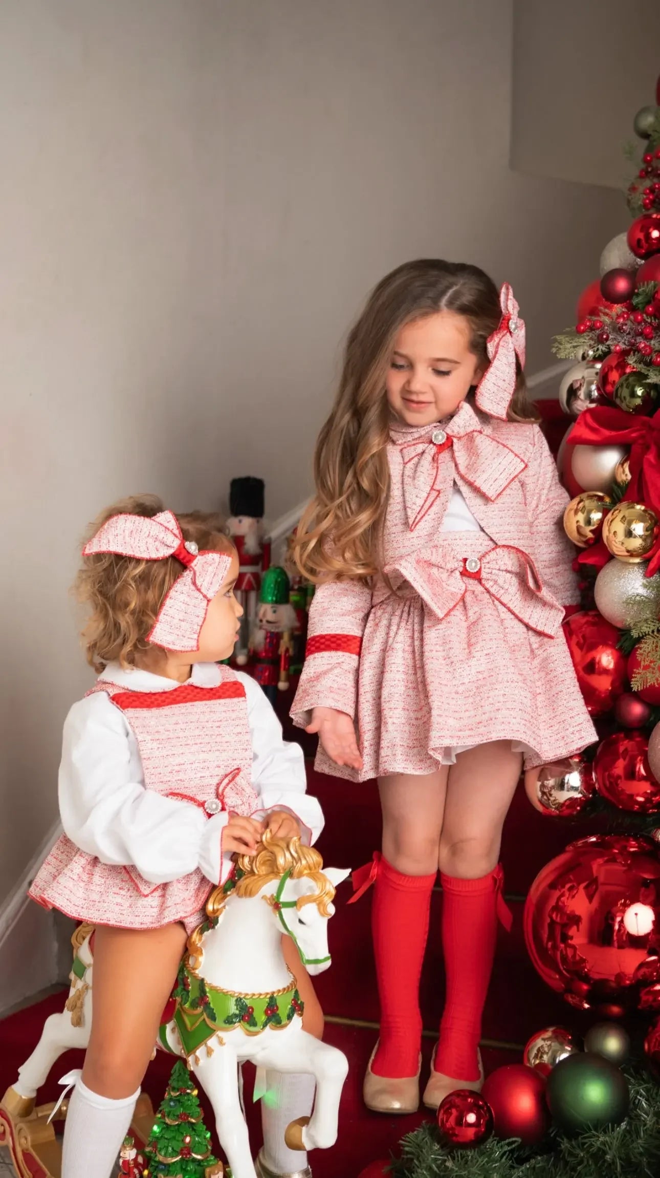 Two young girls in festive red and white outfits near decorated Christmas tree with ornaments and nutcracker toys