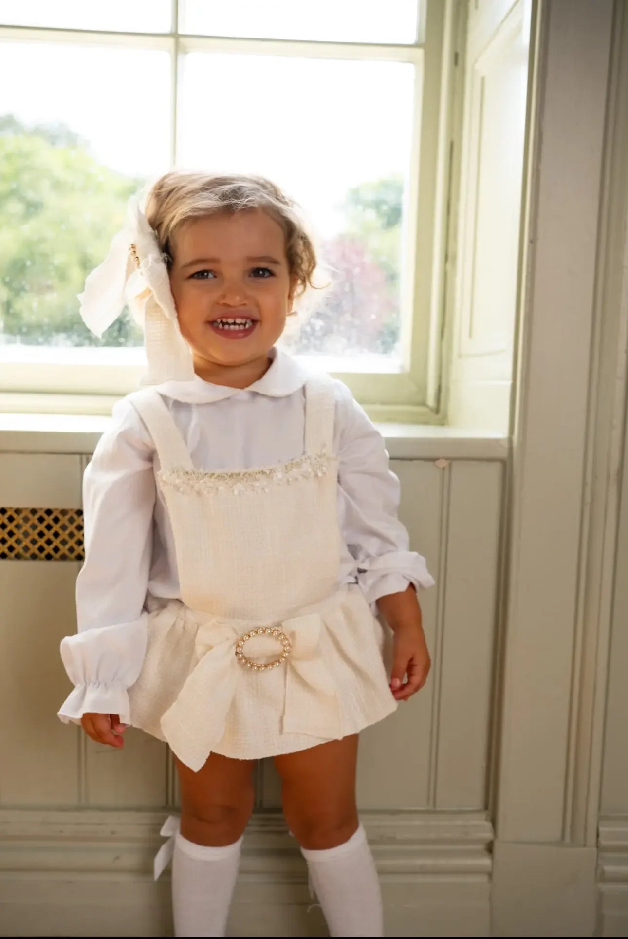 Smiling toddler girl in cream textured dress with bow and white blouse standing by window