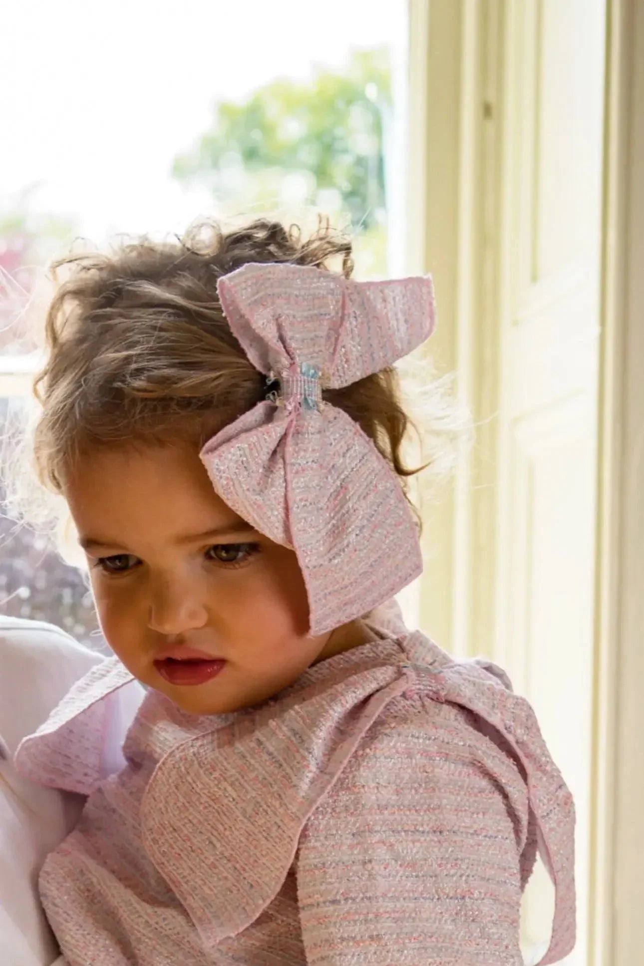 Close-up of toddler girl wearing pink textured dress and large matching bow headband indoors