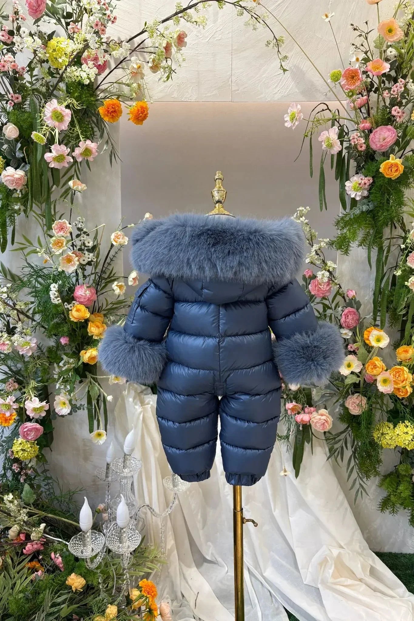 Blue child's puffer snowsuit with fur trim on collar and cuffs displayed on mannequin amidst floral decor