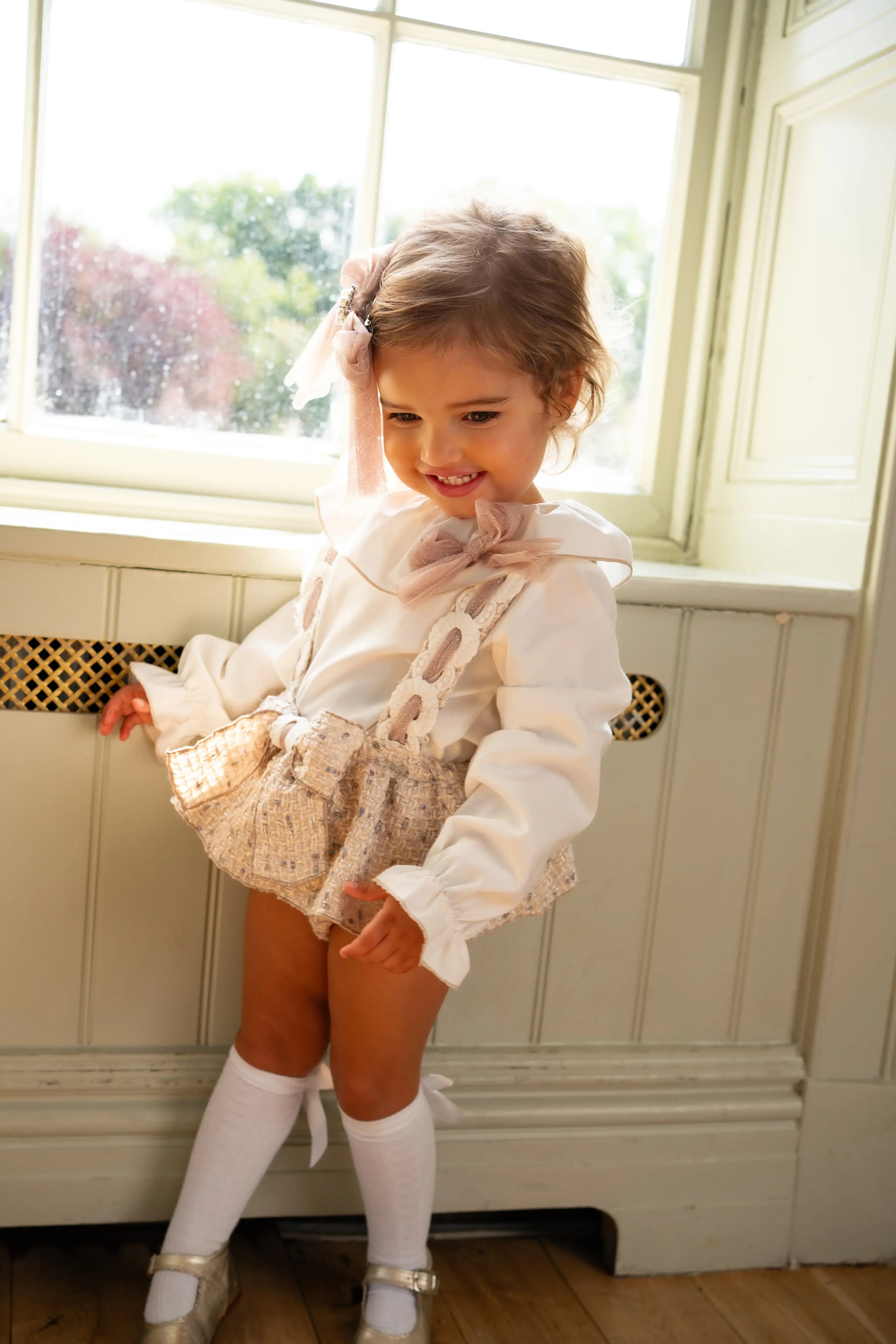 Smiling toddler girl in beige tweed suspender skirt, white blouse, knee-high socks, and bow headband by sunny window