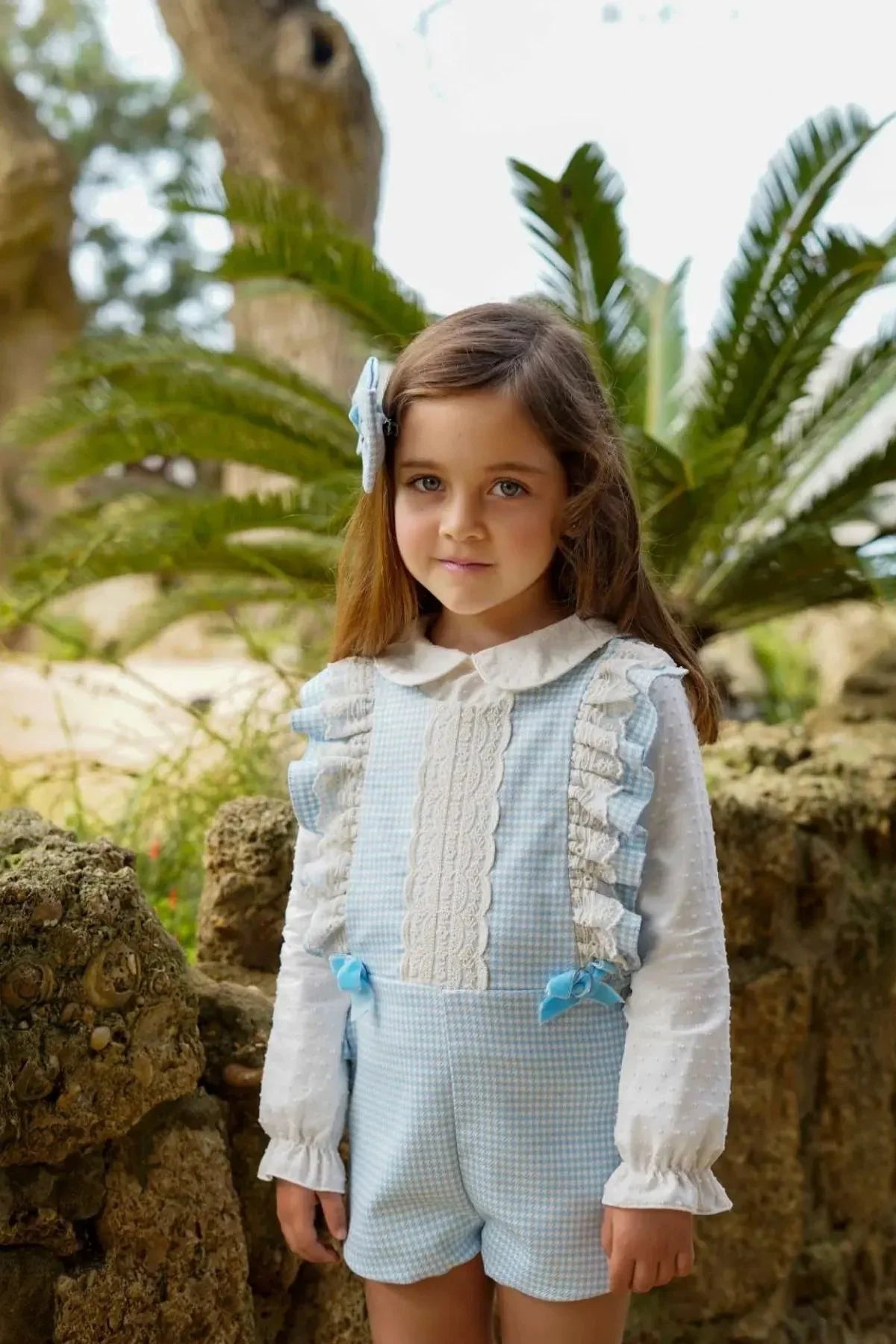 Young girl in blue and white lace romper with ruffles and bow standing outdoors near rocks and palm leaves