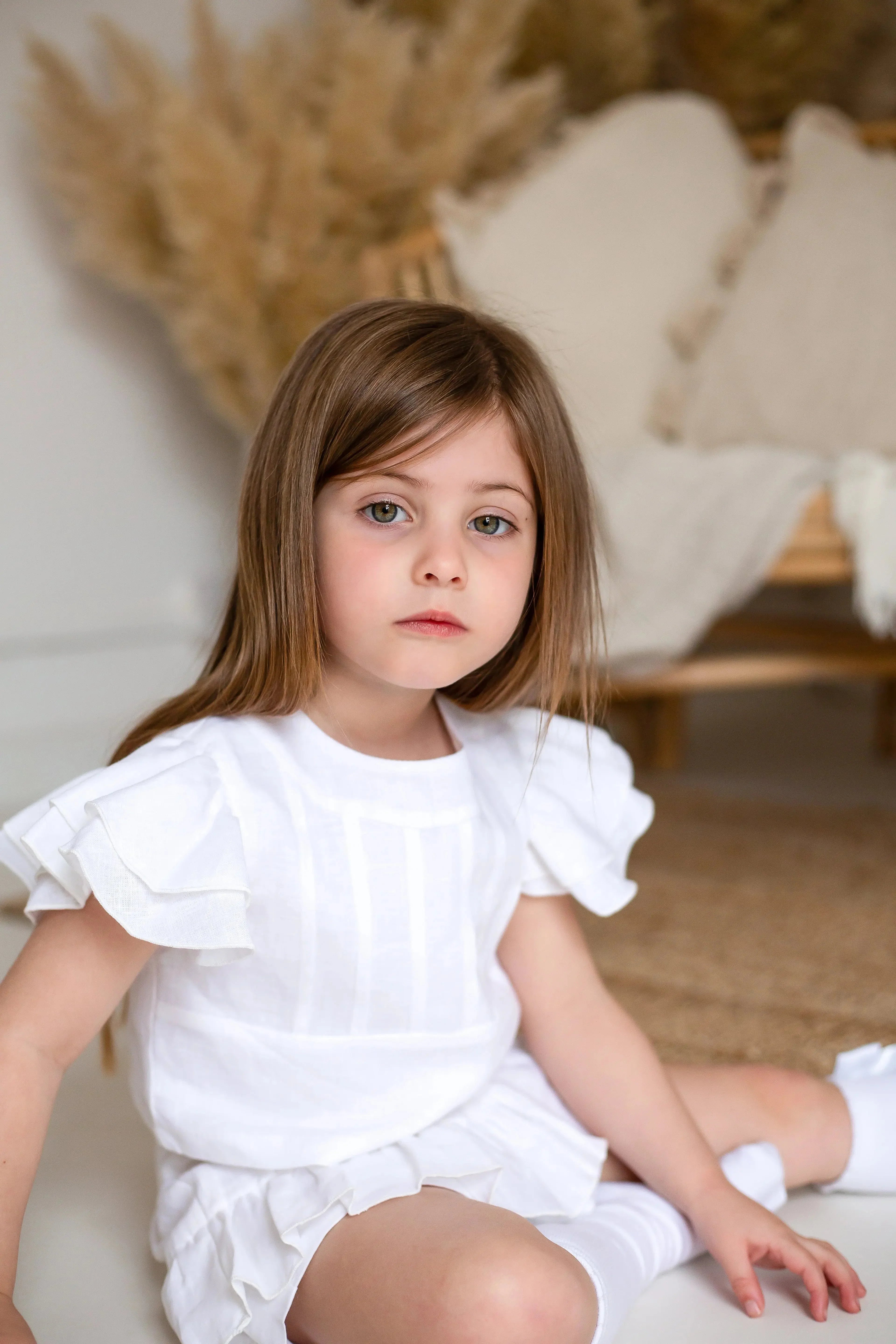 Portrait of young girl with brown hair wearing white ruffled top and shorts sitting indoors