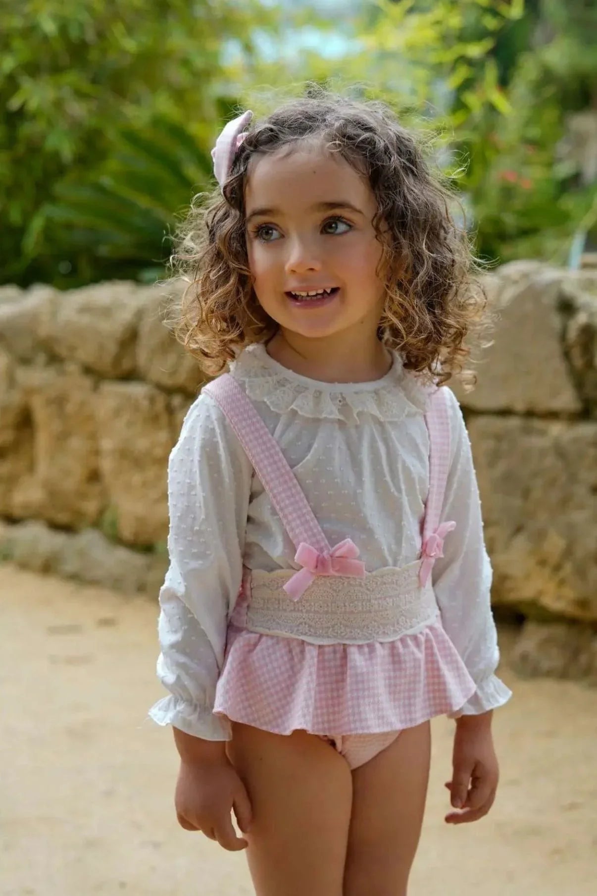 Smiling toddler girl with curly hair wearing white blouse and pink gingham suspender skirt outdoors
