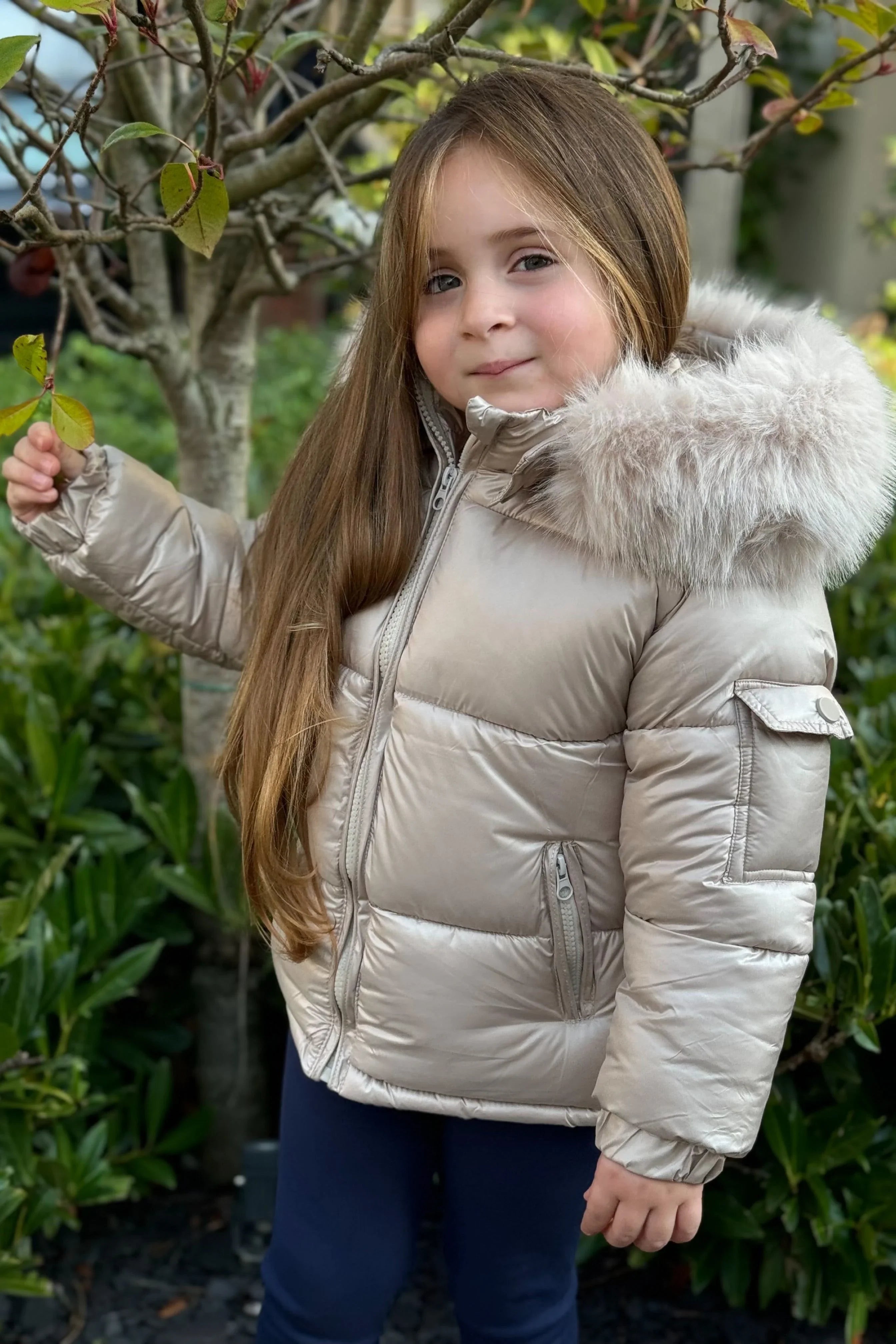 Young girl wearing beige puffer jacket with fur hood standing outdoors near tree and green bushes