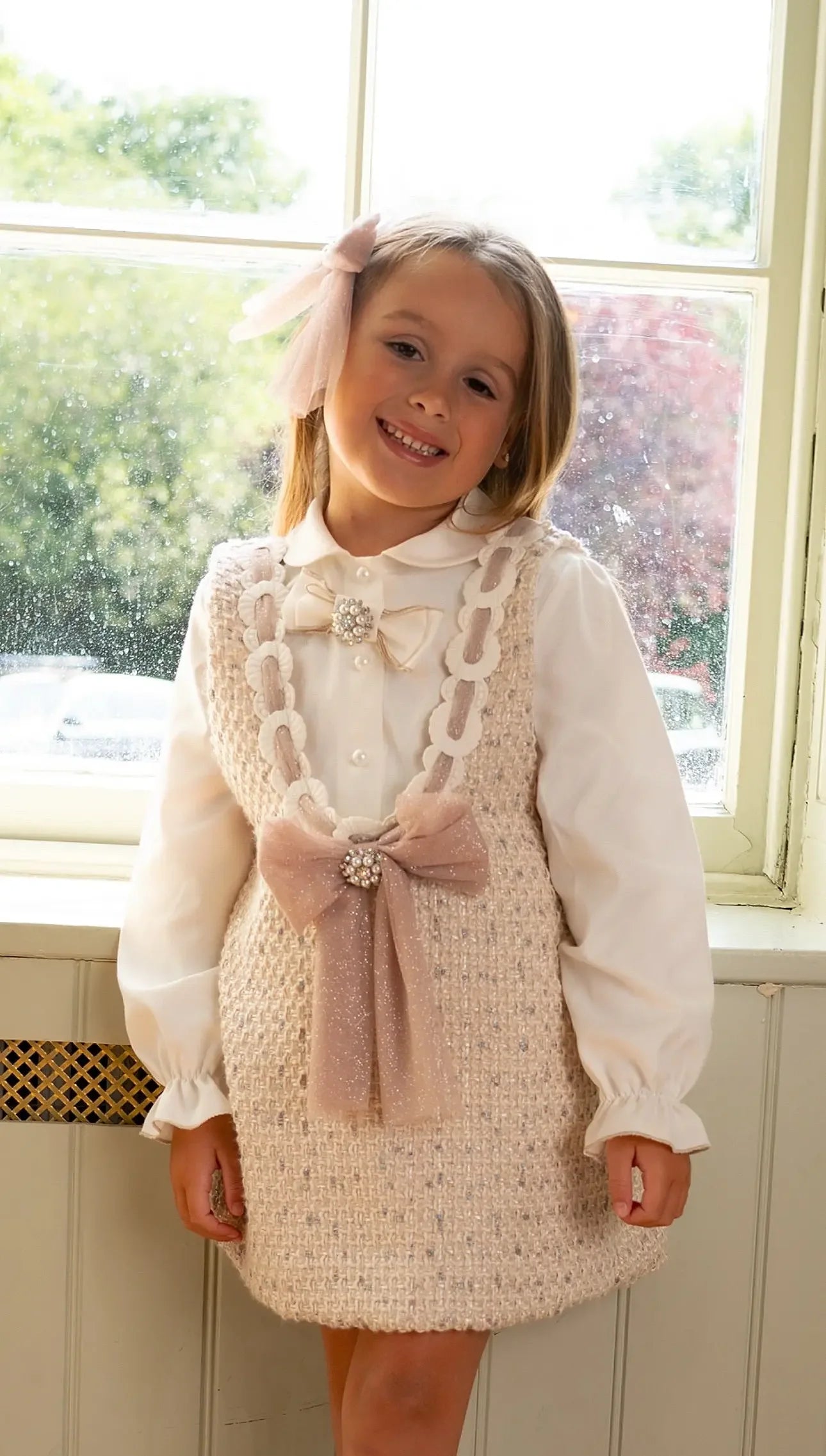 Smiling girl in white blouse and beige textured dress with pearl bows standing by window