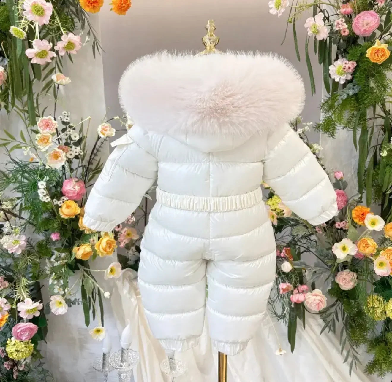 White baby snowsuit with furry hood displayed on mannequin surrounded by colorful flowers