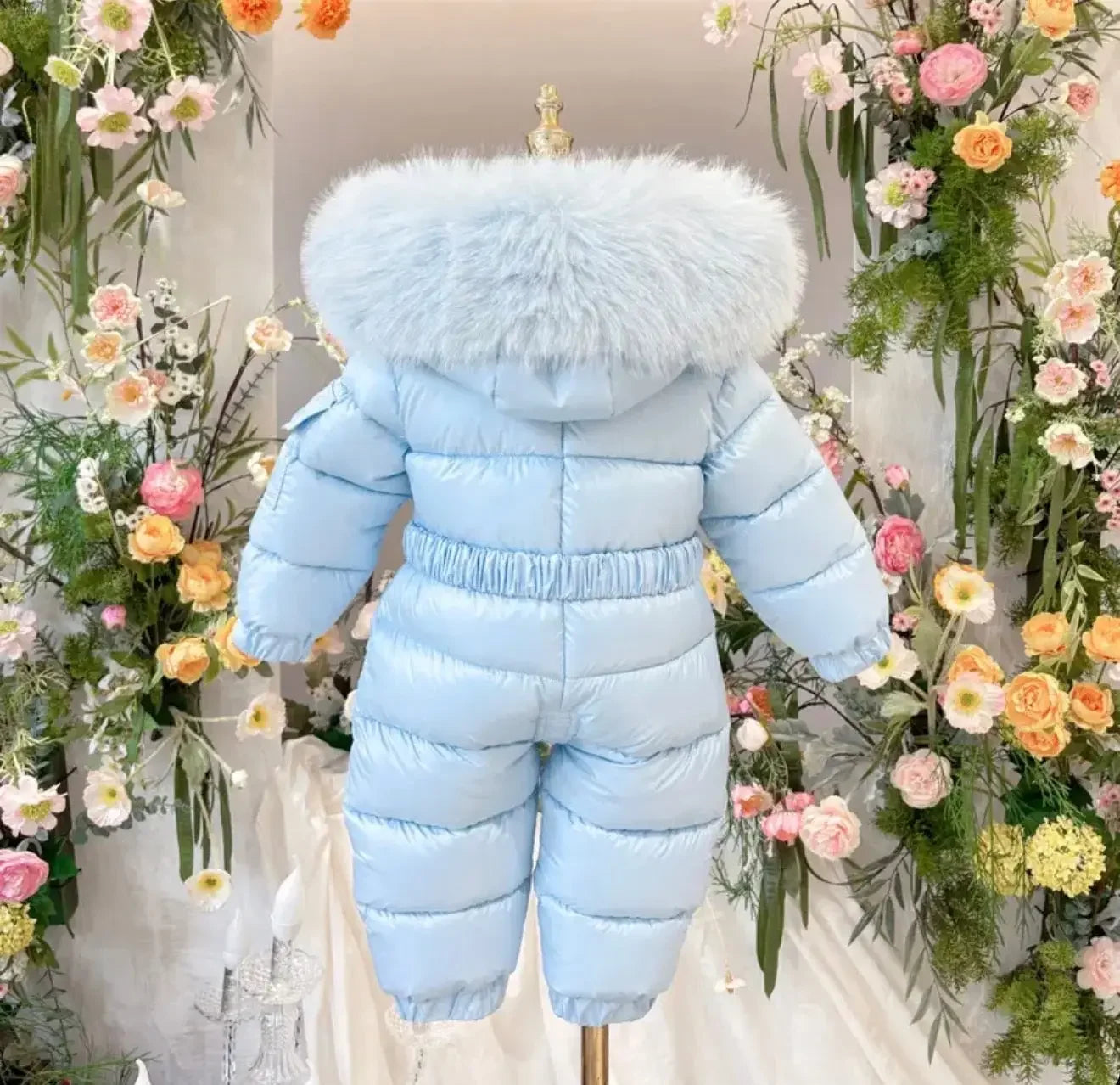 Light blue puffer toddler snowsuit with fluffy hood displayed on mannequin surrounded by colorful flowers