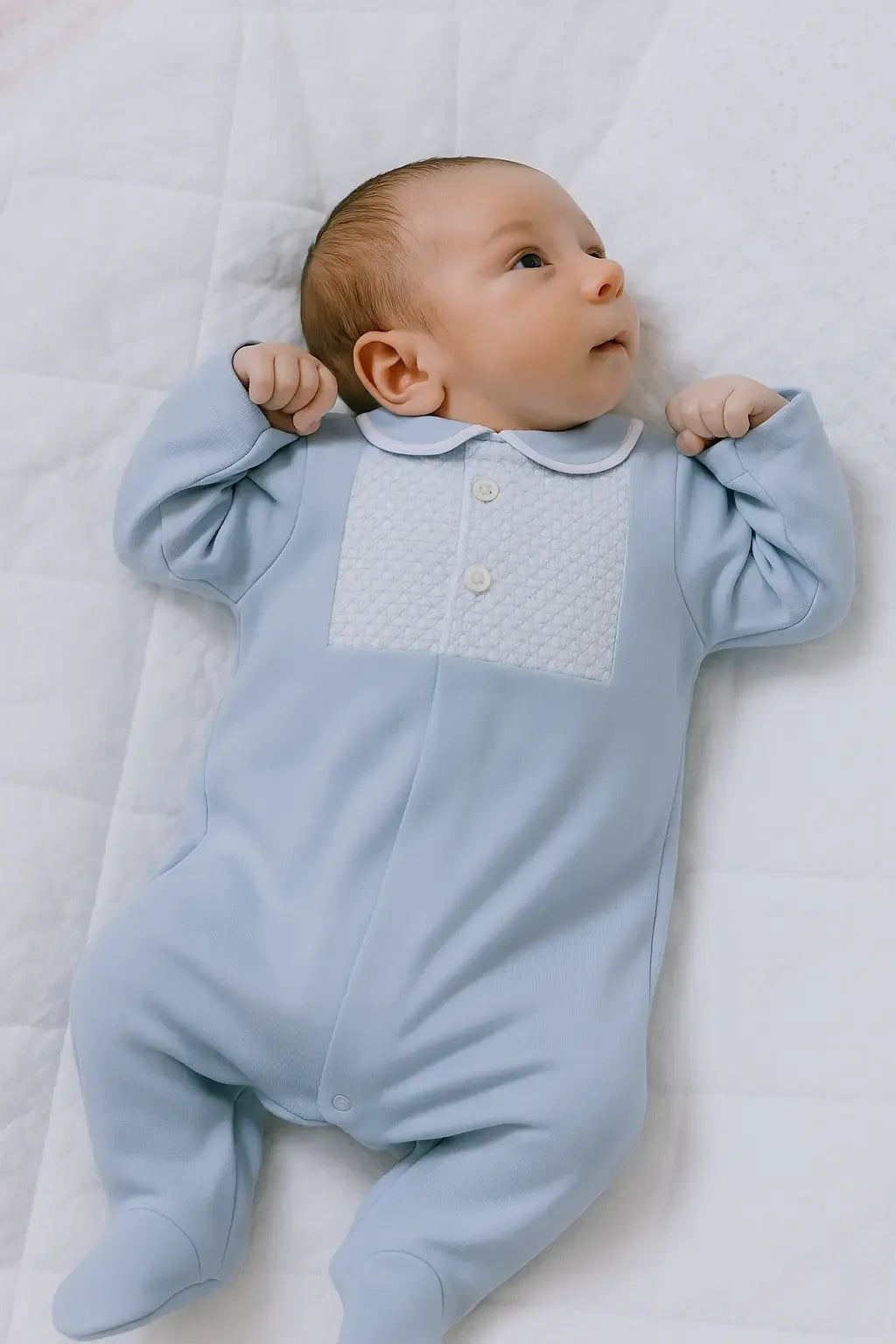Newborn baby lying on white quilted blanket wearing light blue onesie with textured chest panel