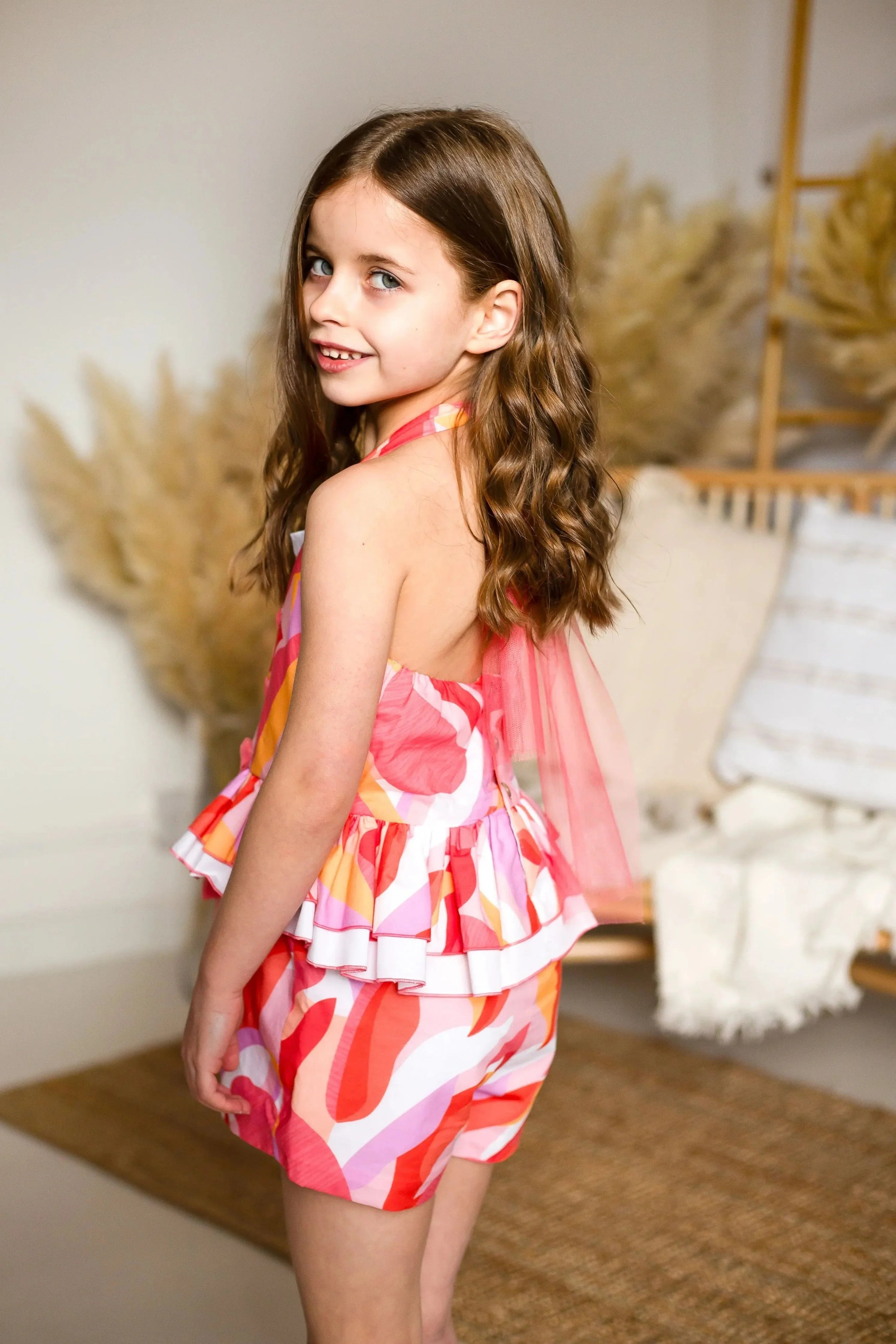 Smiling young girl with long brown hair wearing colorful summer outfit with ruffled top and shorts indoors