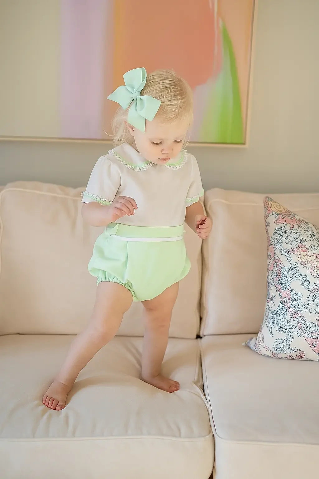 Toddler girl with blonde hair wearing mint green bow and romper standing on cream sofa with decorative pillow