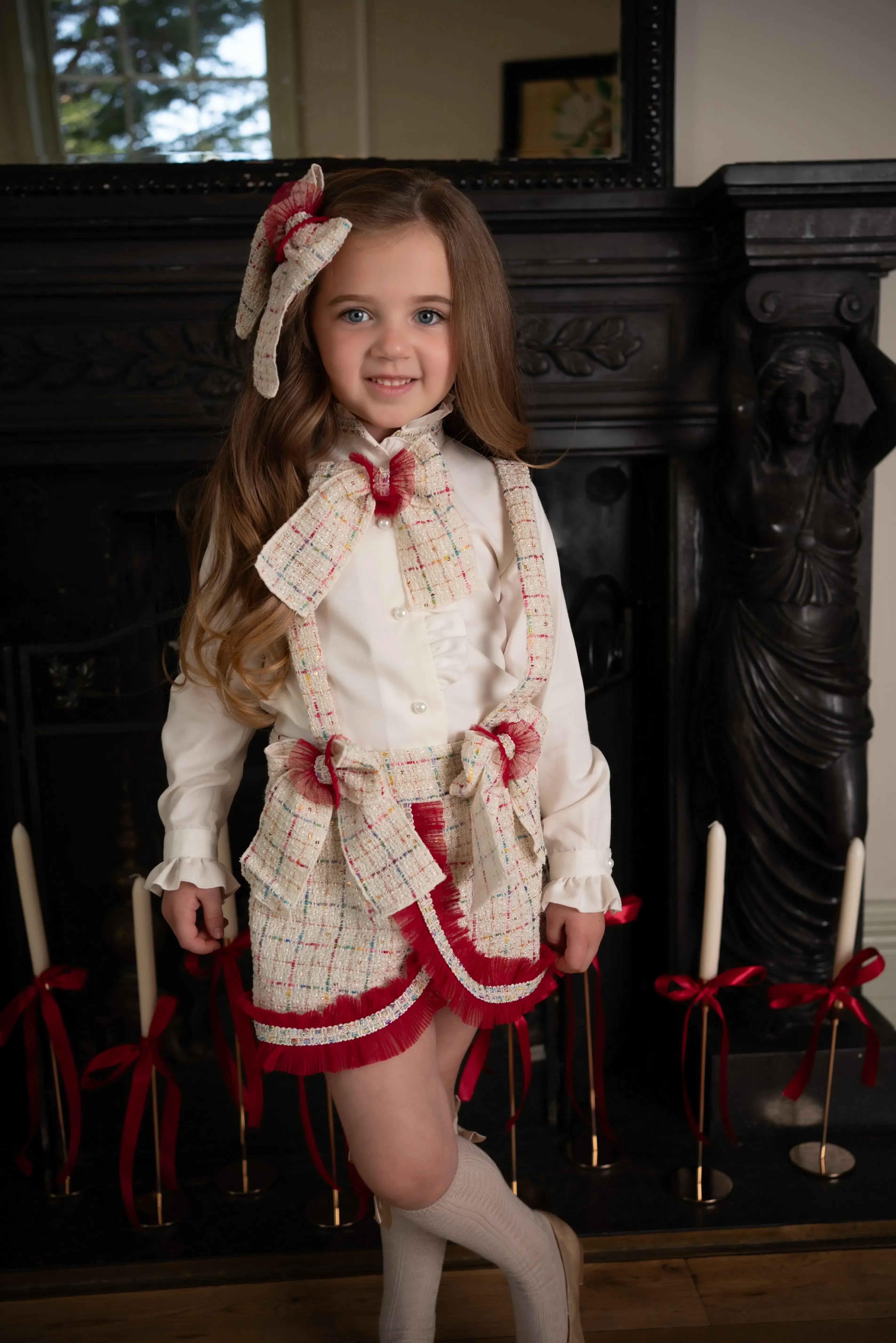 Young girl in white blouse and plaid skirt with red accents standing by black ornate fireplace with candles