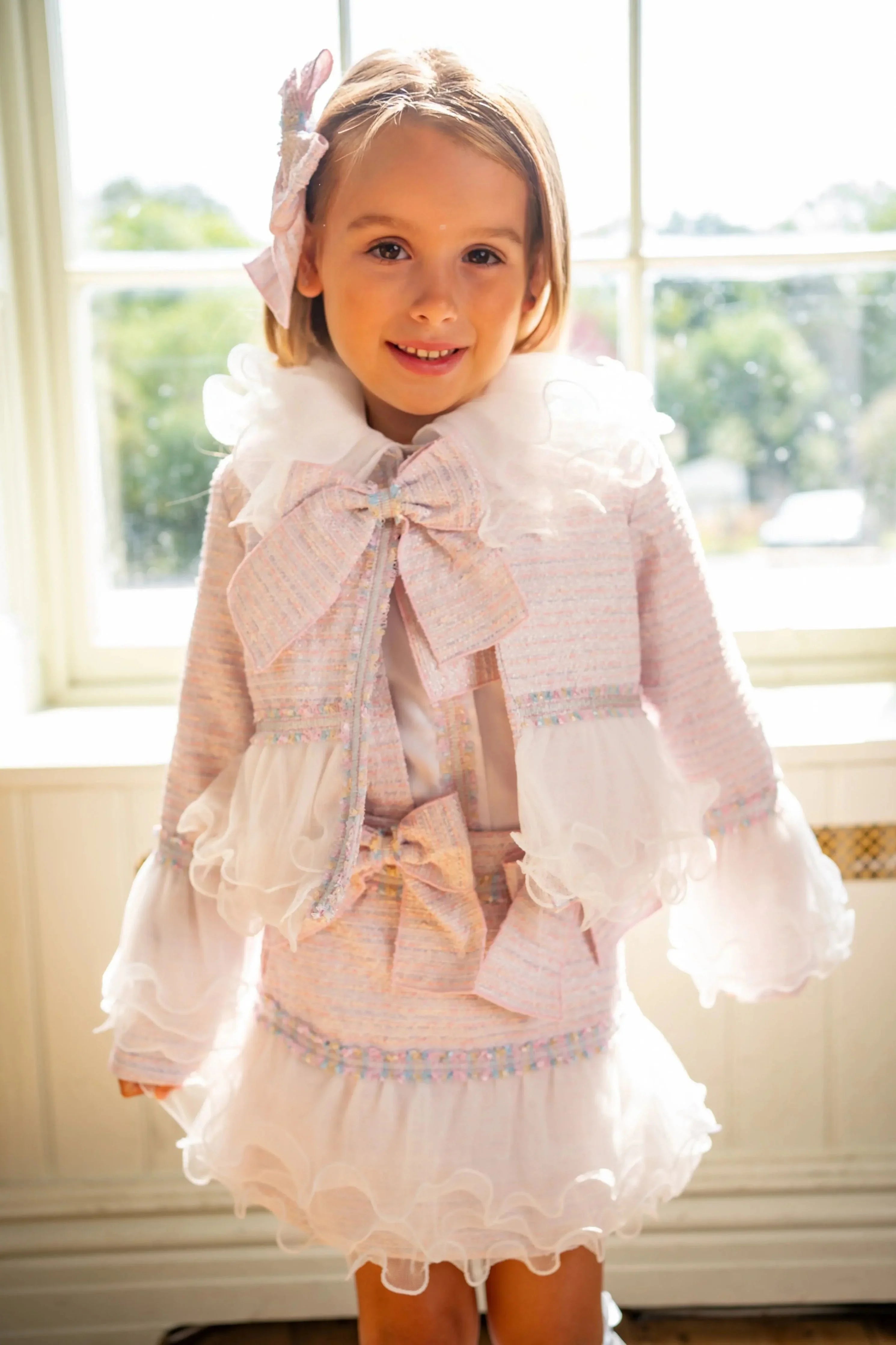 Smiling girl in pastel pink ruffled dress and jacket with bows, standing by sunlit window