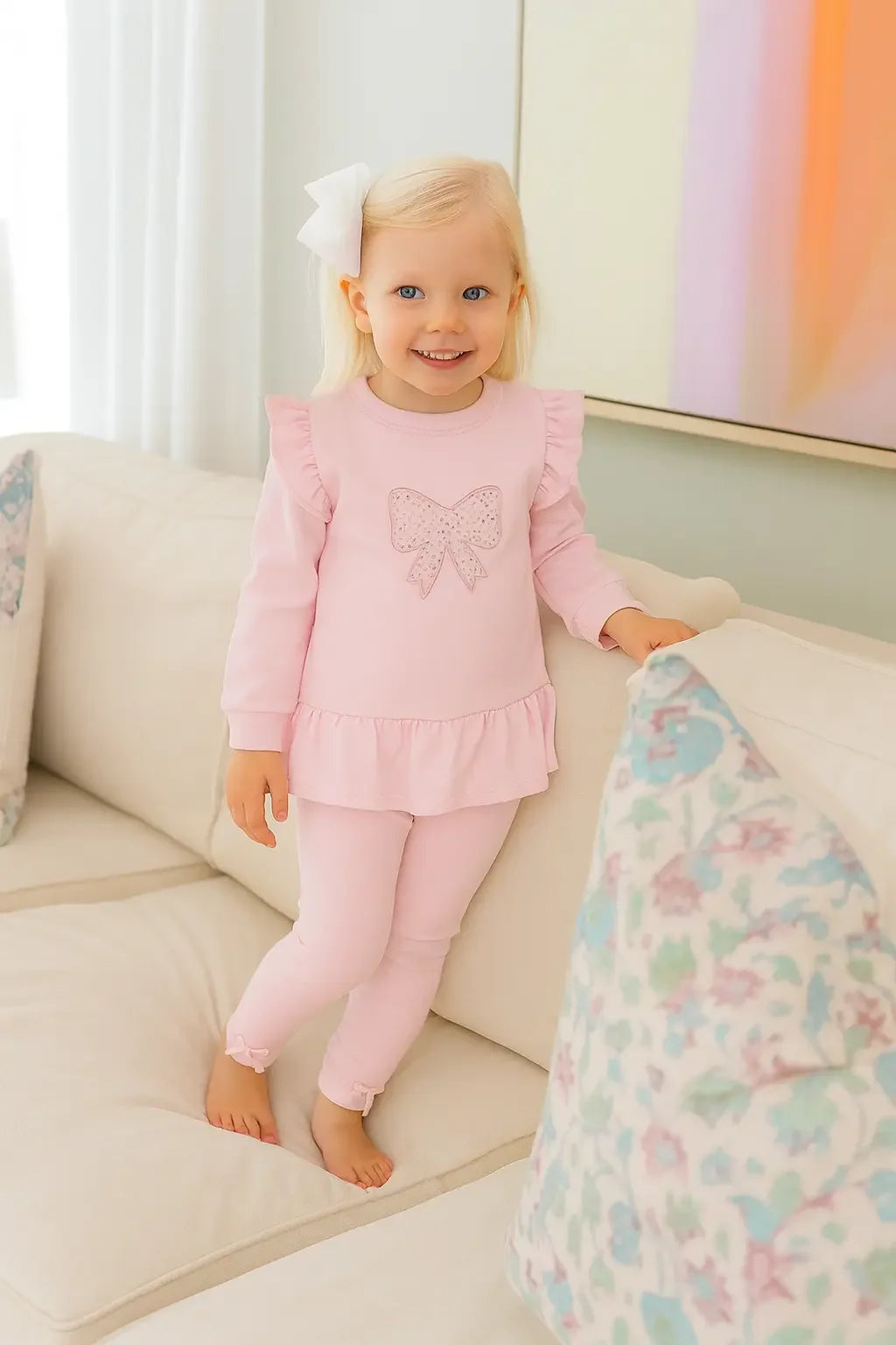 Smiling toddler girl in pink leggings and ruffled top with bow detail standing on beige couch indoors