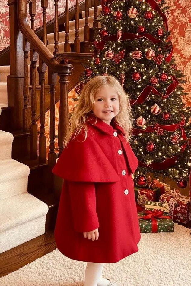Little girl in a red coat standing near decorated Christmas tree and wooden staircase