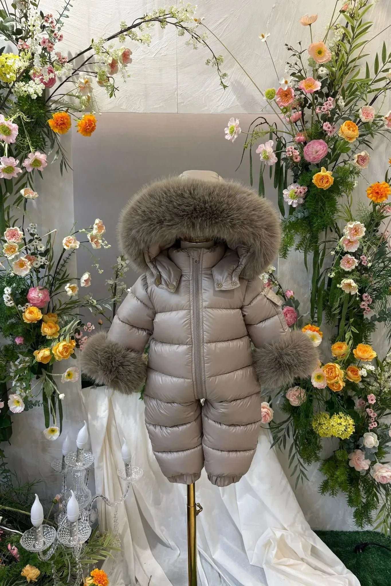 Beige baby winter snowsuit with fur hood and cuffs displayed on a mannequin amid colorful flowers