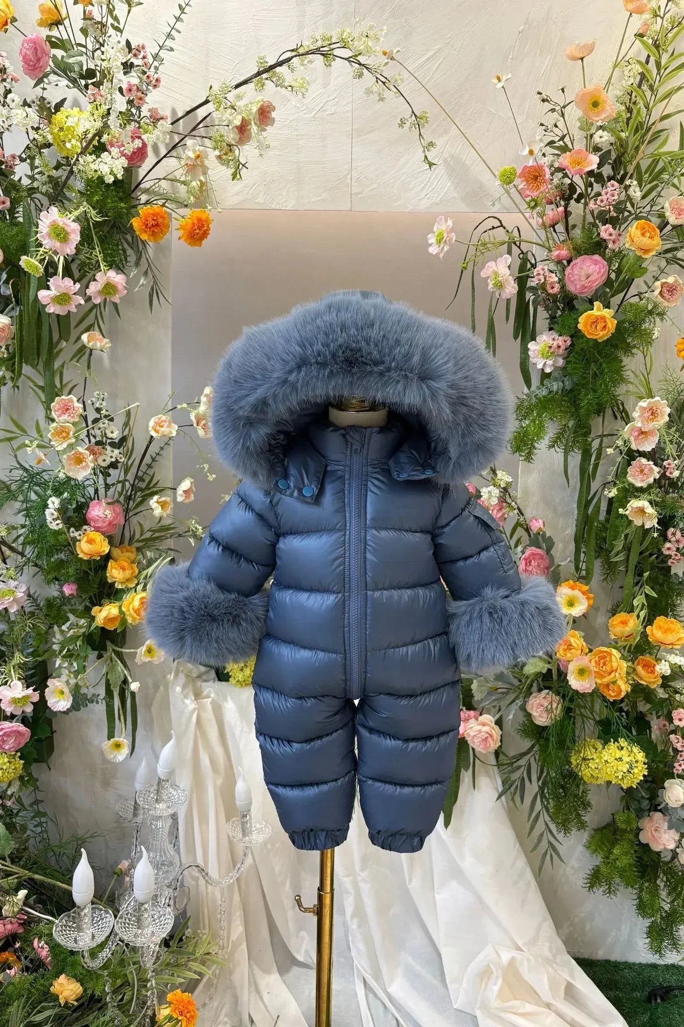 Blue baby snowsuit with large fur hood and cuffs on mannequin surrounded by colorful floral arrangements