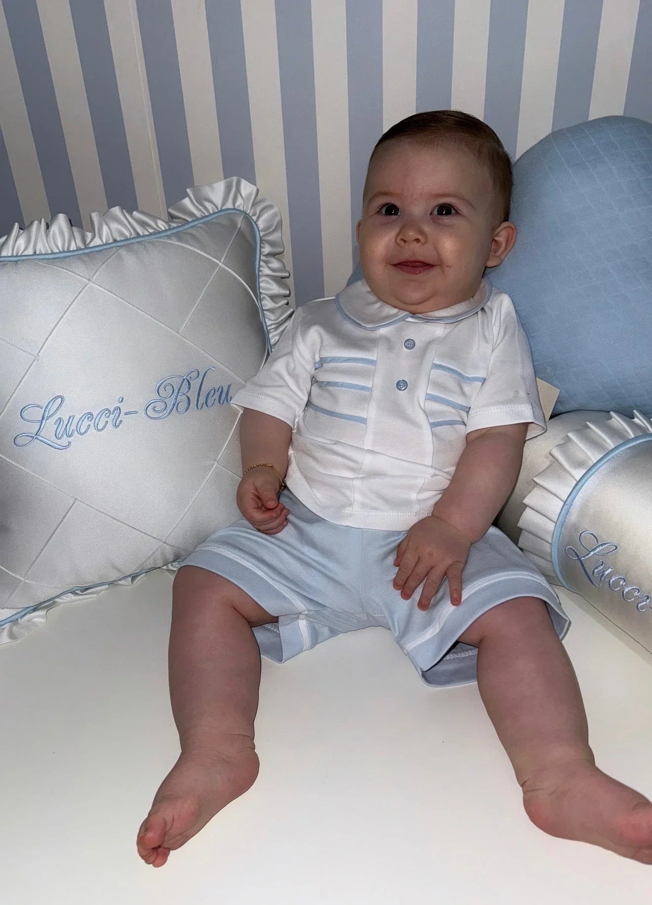 Smiling baby boy in white and light blue outfit sitting with Lucci-Bleu embroidered pillows
