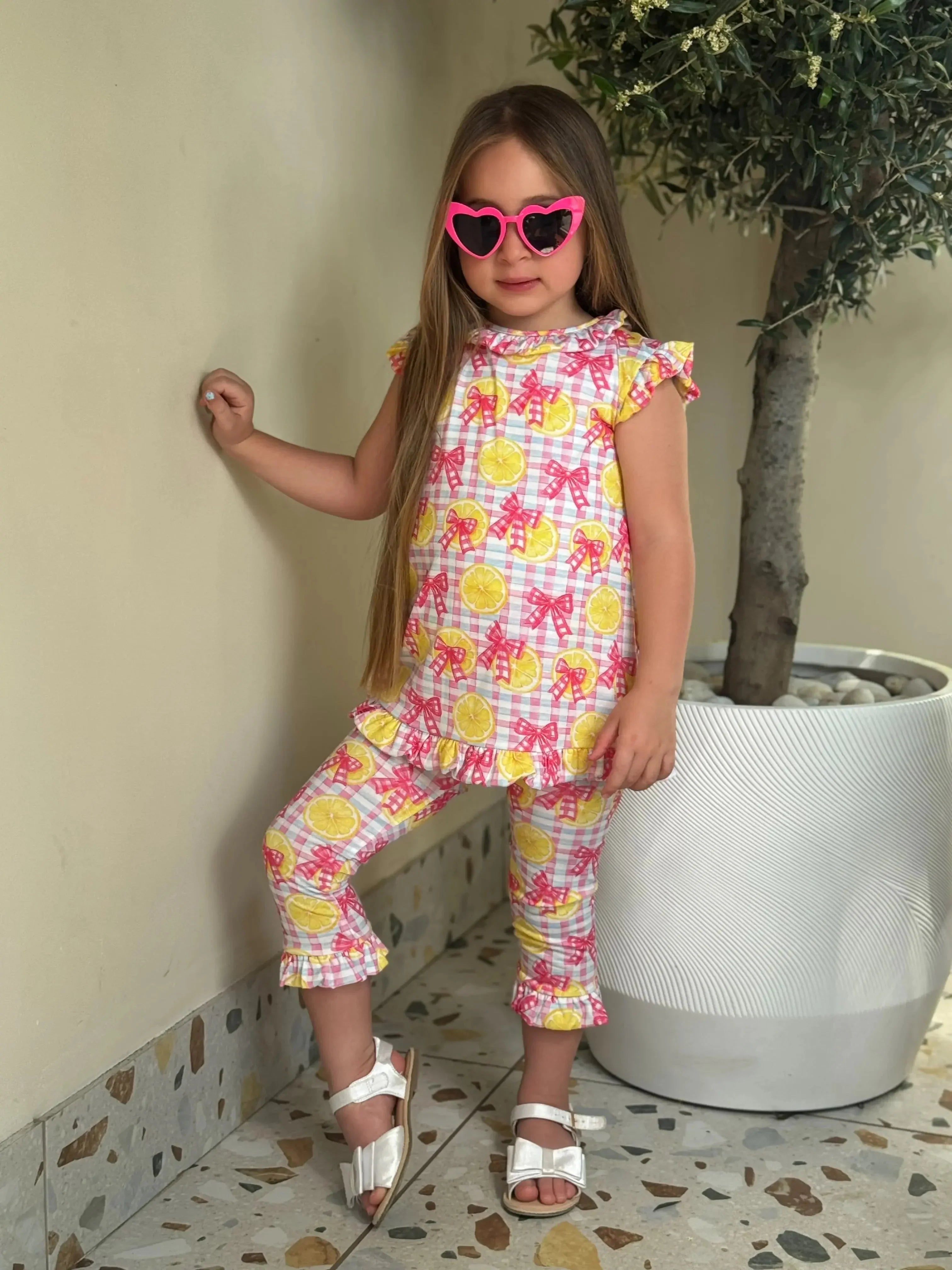 young girl wearing matching lemon and pink bow print outfit with heart-shaped pink sunglasses indoors