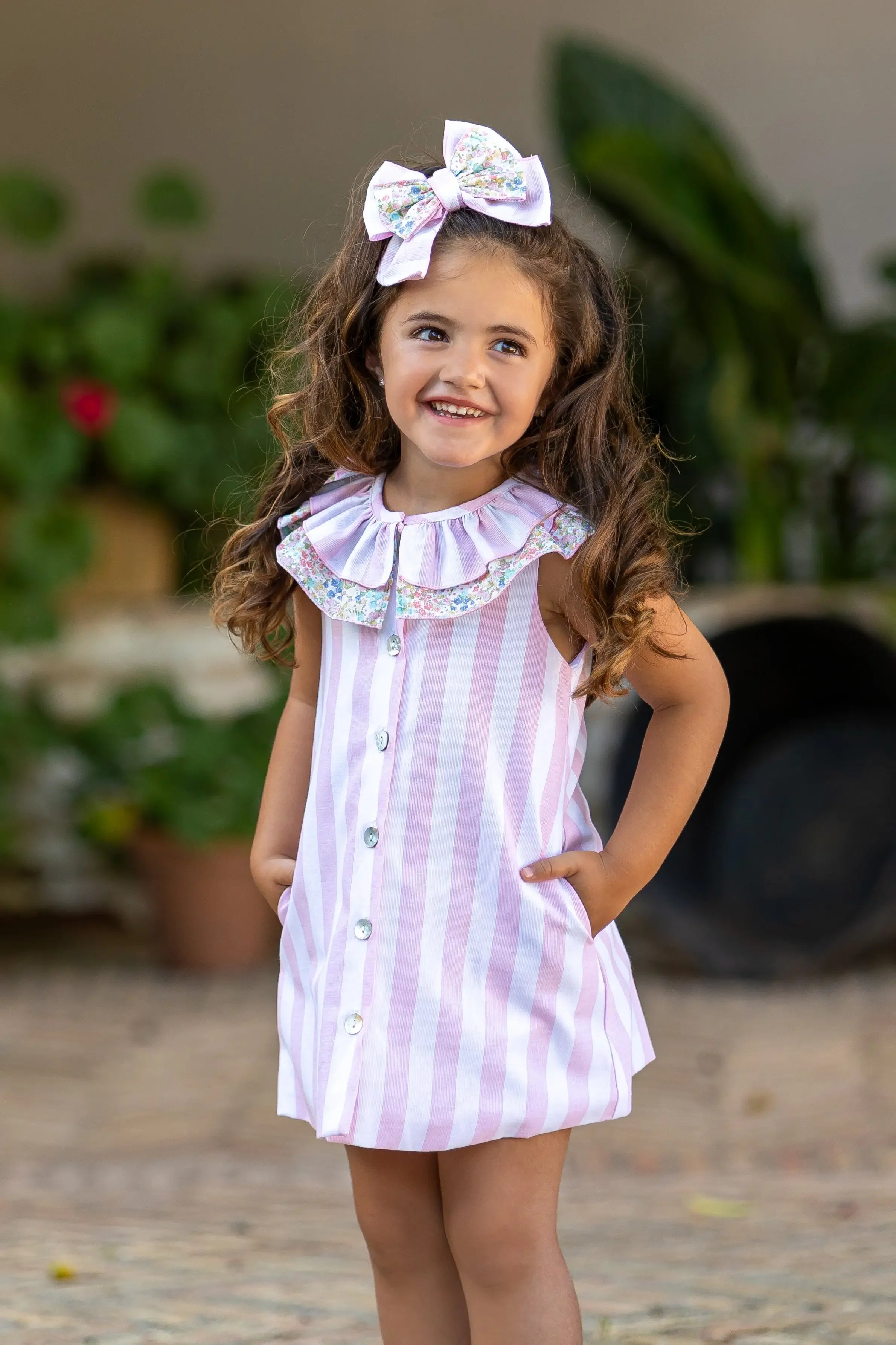 Smiling little girl with long curly hair wearing pink and white striped dress and floral bow outdoors
