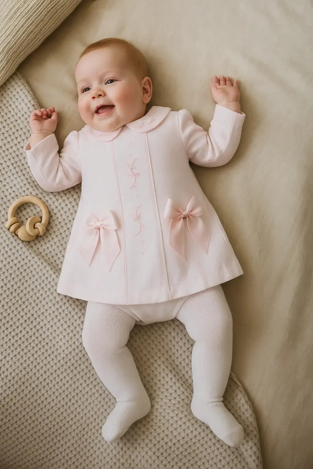 Smiling baby in pink dress with bows and white tights lying on beige textured blanket with wooden rattle