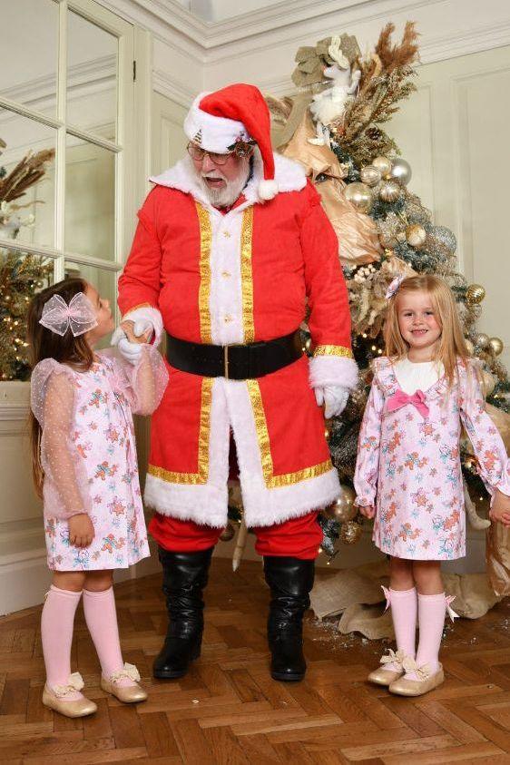 Santa Claus in red suit holding hands with two girls in pink floral dresses and knee-high socks near Christmas tree