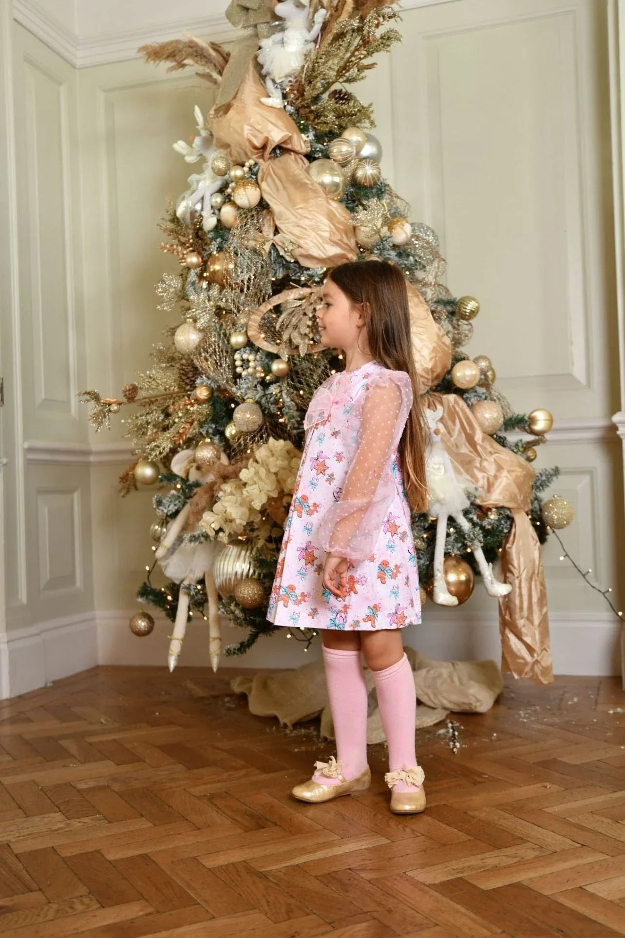 Little girl in a floral gingerbread dress with organza sleeves and pink knee socks standing by a gold-decorated Christmas tree