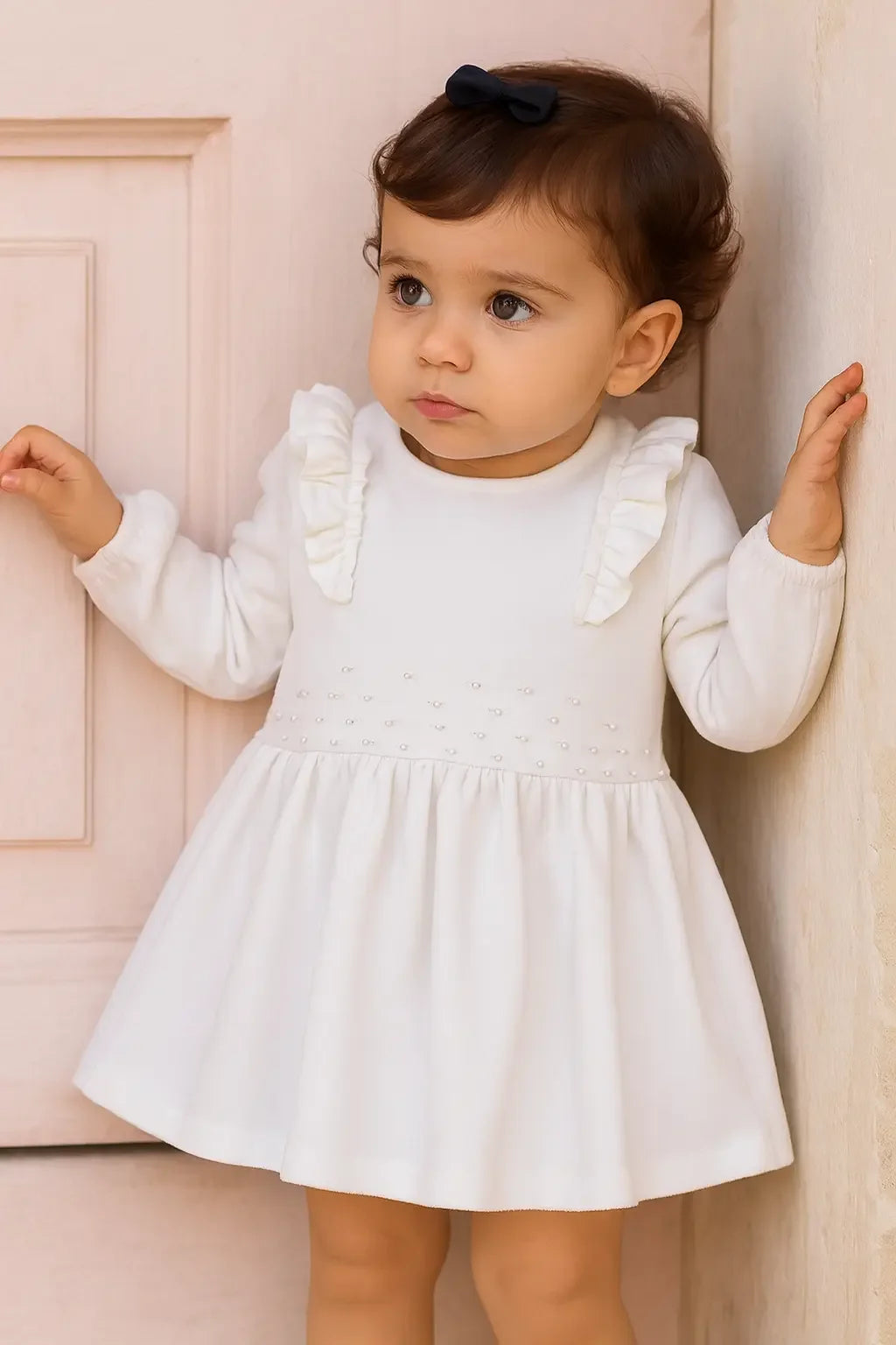 Toddler girl wearing a cream pearl dress with ruffles, standing by a light pink door and wall