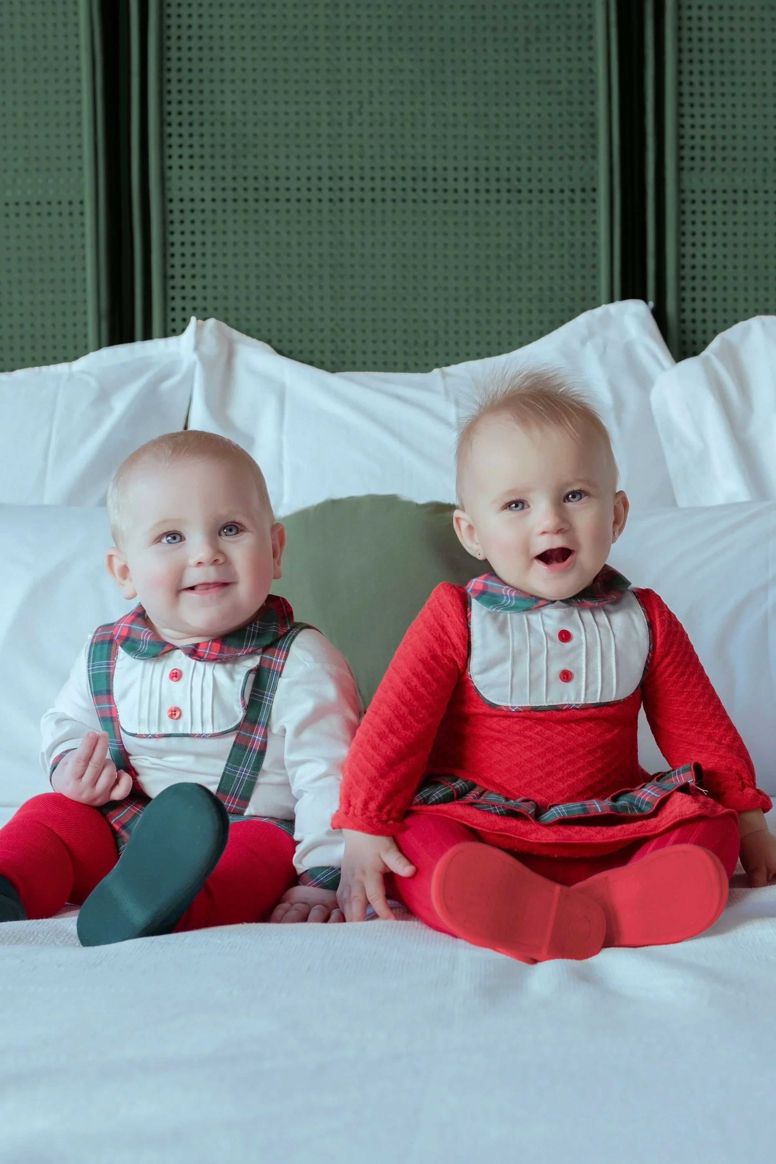 Two smiling babies in festive red and plaid holiday outfits sitting on a white bed with a green cushioned headboard