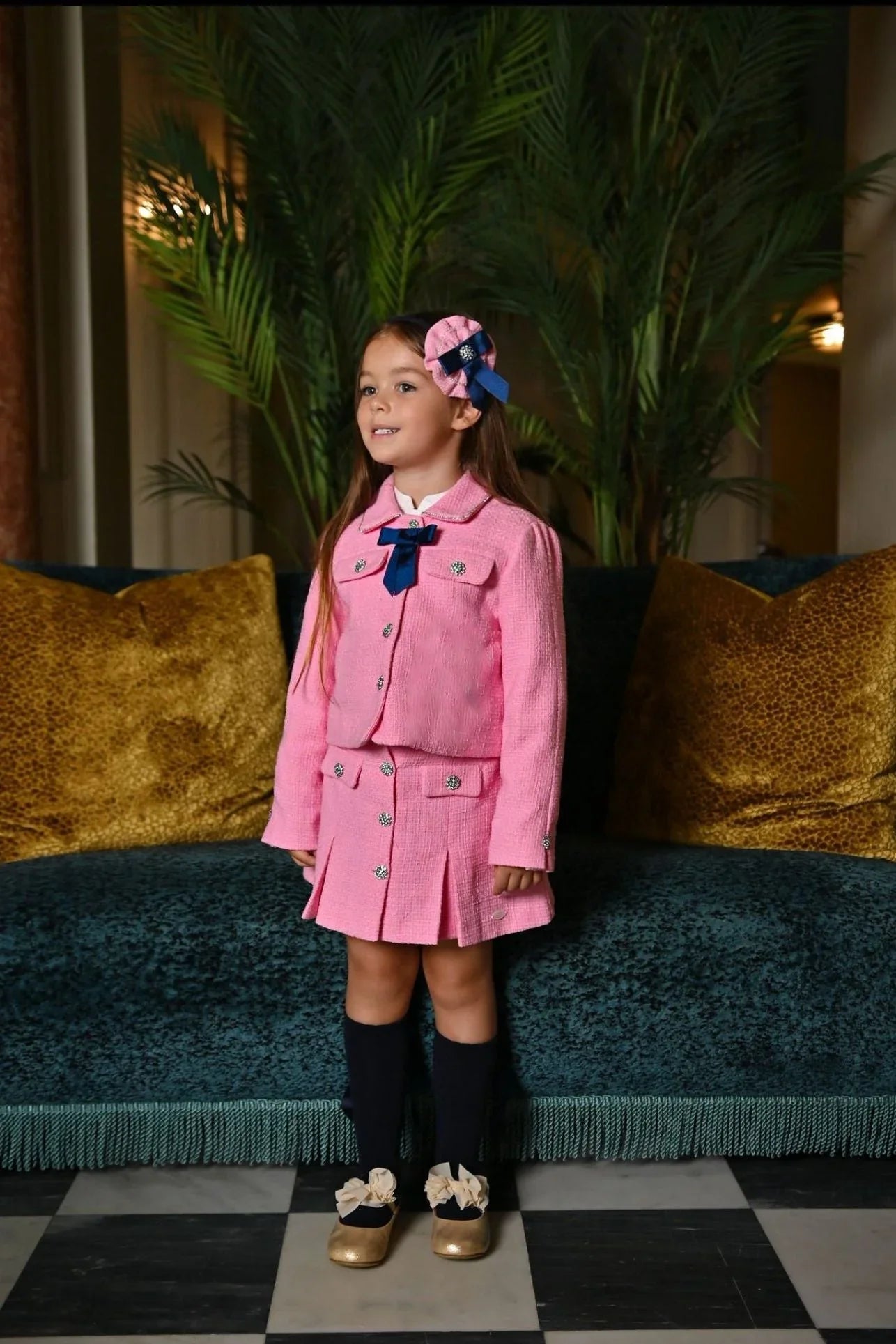 Young girl wearing a pink tweed skirt suit with navy bow and matching headpiece, standing indoors