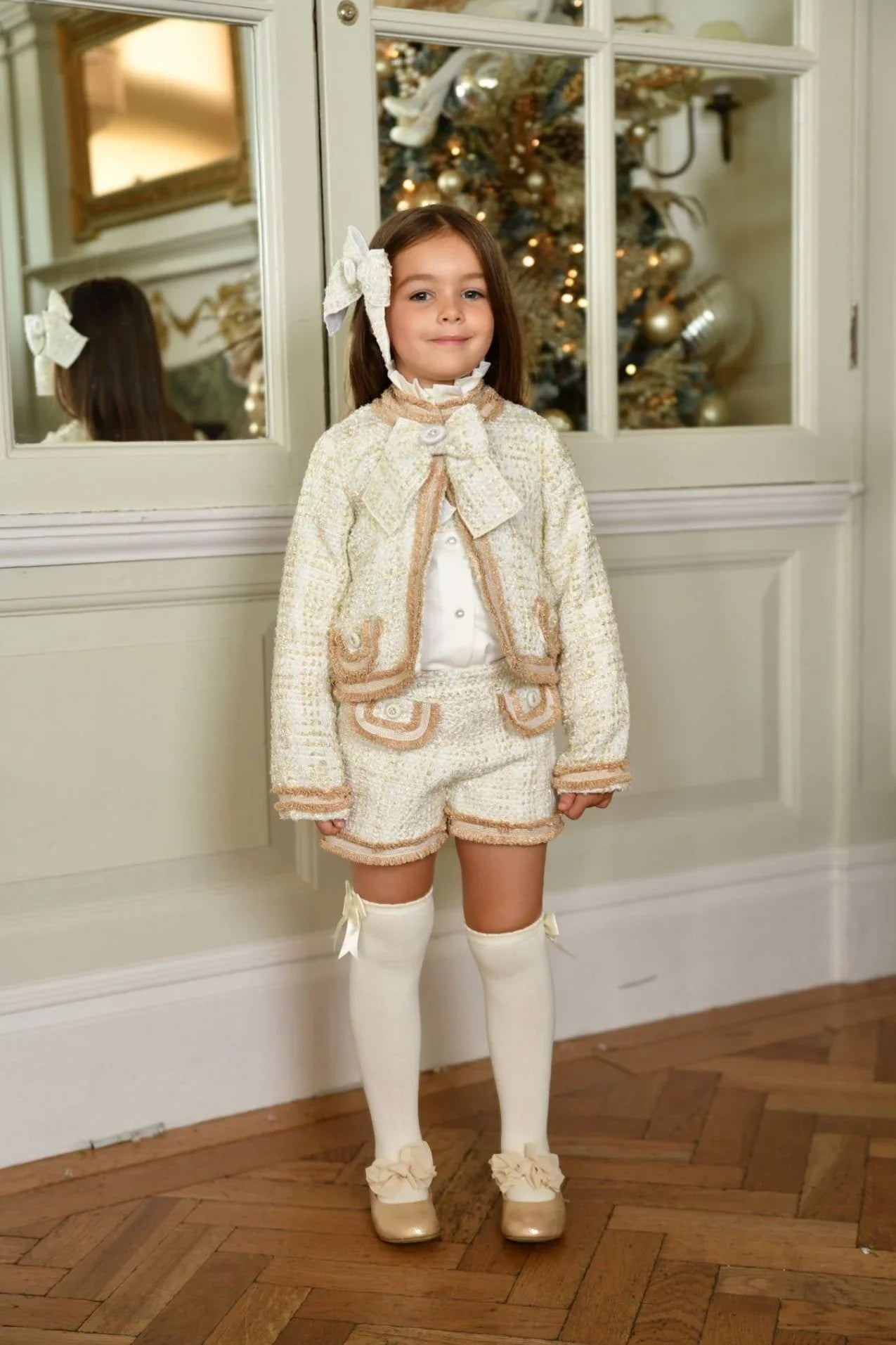 Young girl in cream tweed Chanel blazer and shorts set, knee-high socks, standing indoors near mirror and decorated Christmas tree