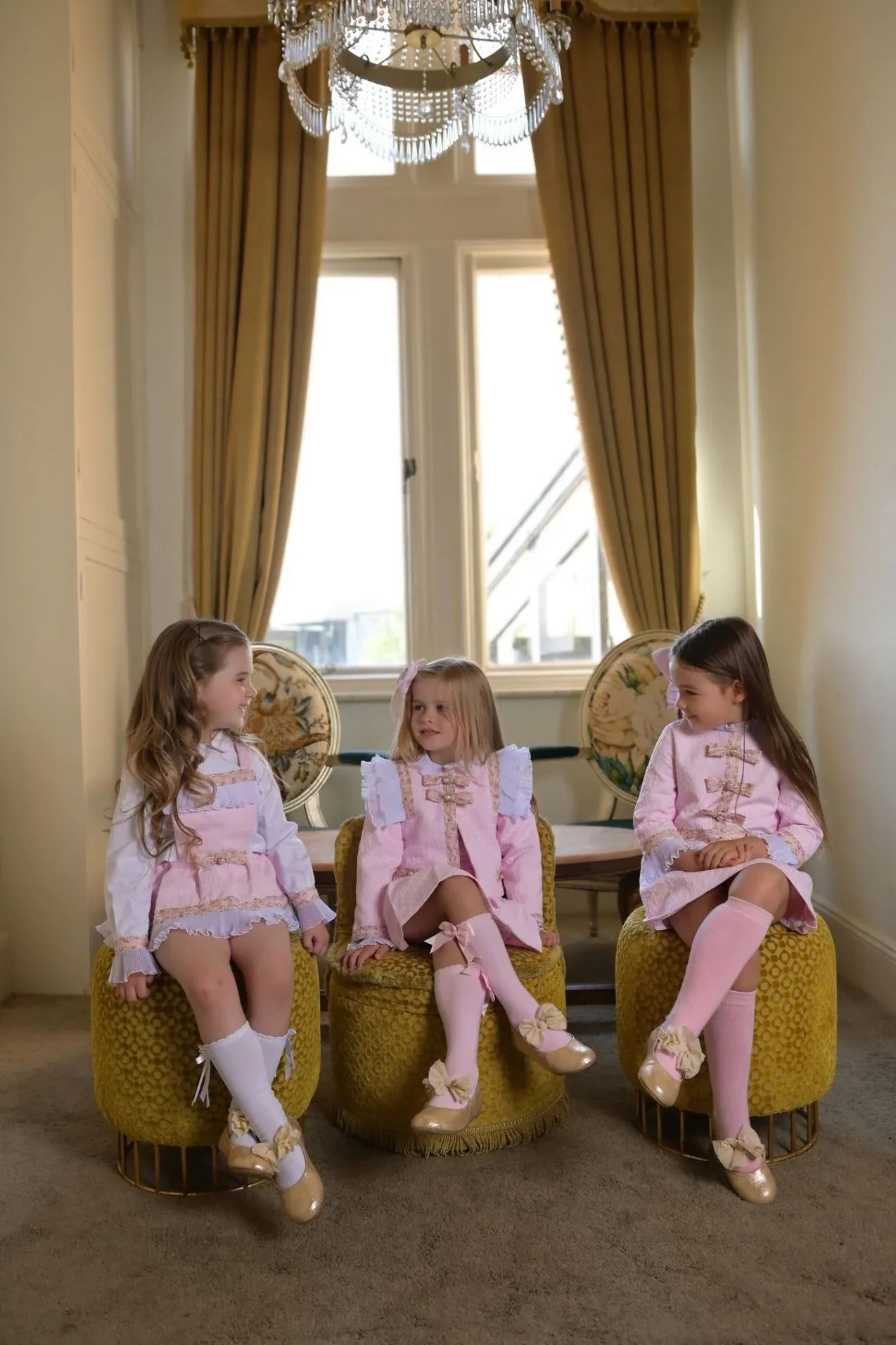 Three young girls in pink vintage-style dresses with bows sitting on yellow ottomans in elegant room