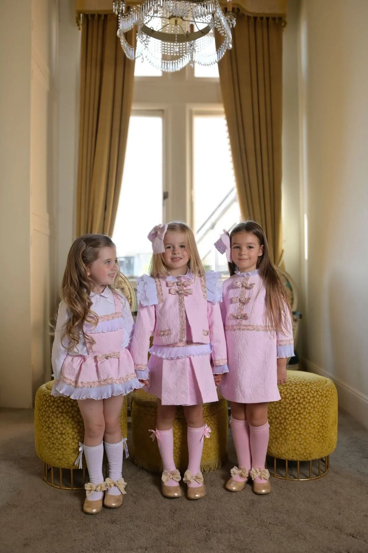 Three young girls in pink damask dresses standing in a room with mustard yellow poufs and gold curtains