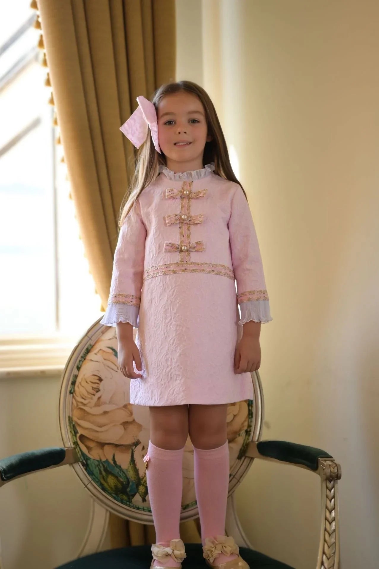 Young girl wearing a pink damask dress with bows, knee-high socks, and a large hair bow standing indoors