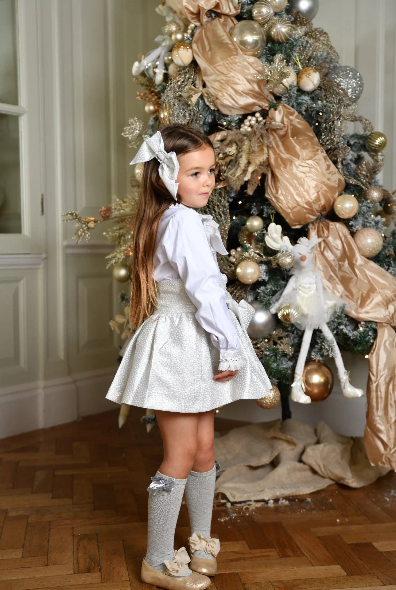 Girl in silver croc hair bow, white blouse, and silver skirt standing by elegant Christmas tree