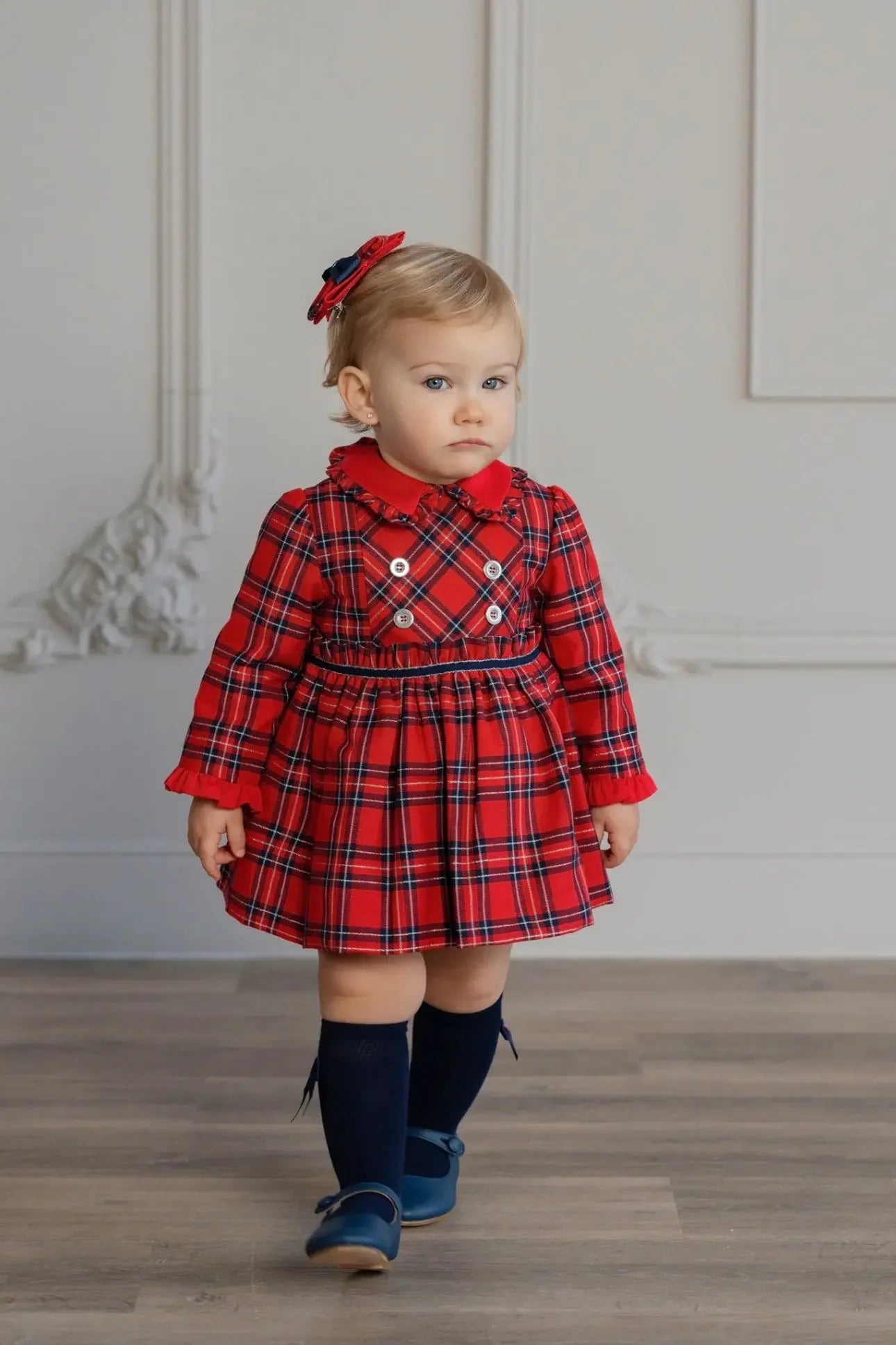 Toddler girl in red tartan dress with long sleeves, navy socks, and flower hair clip standing indoors