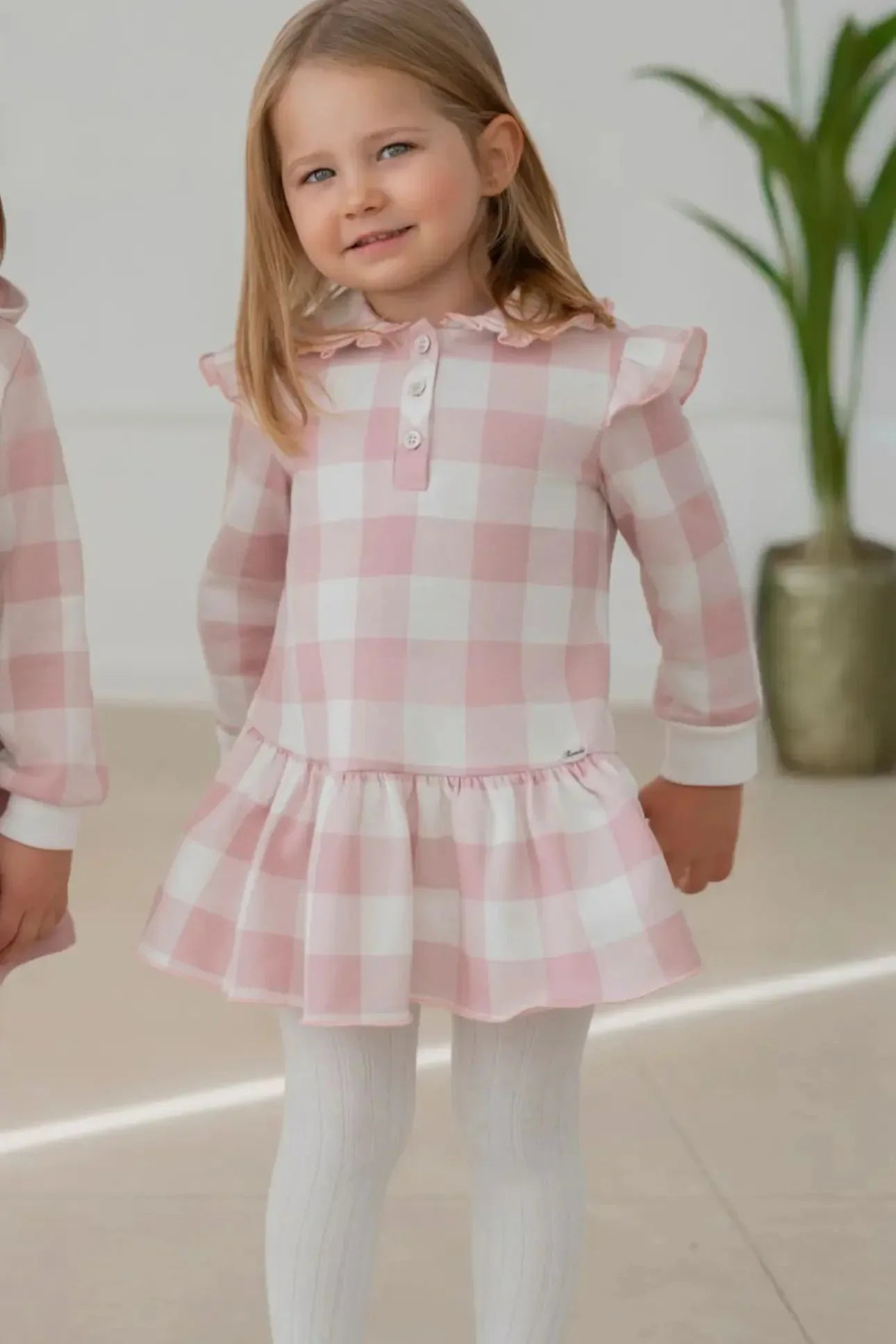 Smiling girl wearing pink and white checkered dress with ruffle collar and white tights indoors