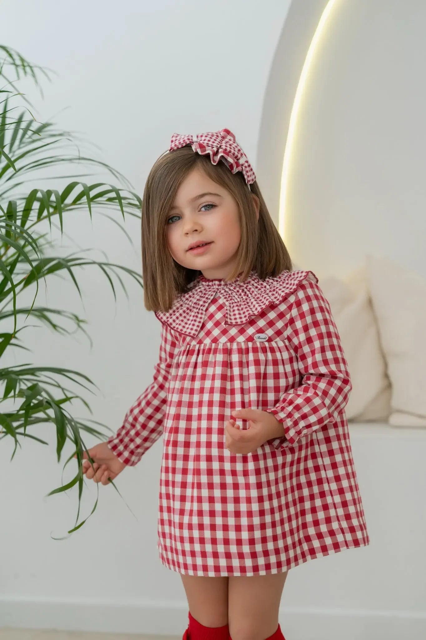 Little girl in red and white checkered dress and matching headband holding a plant indoors