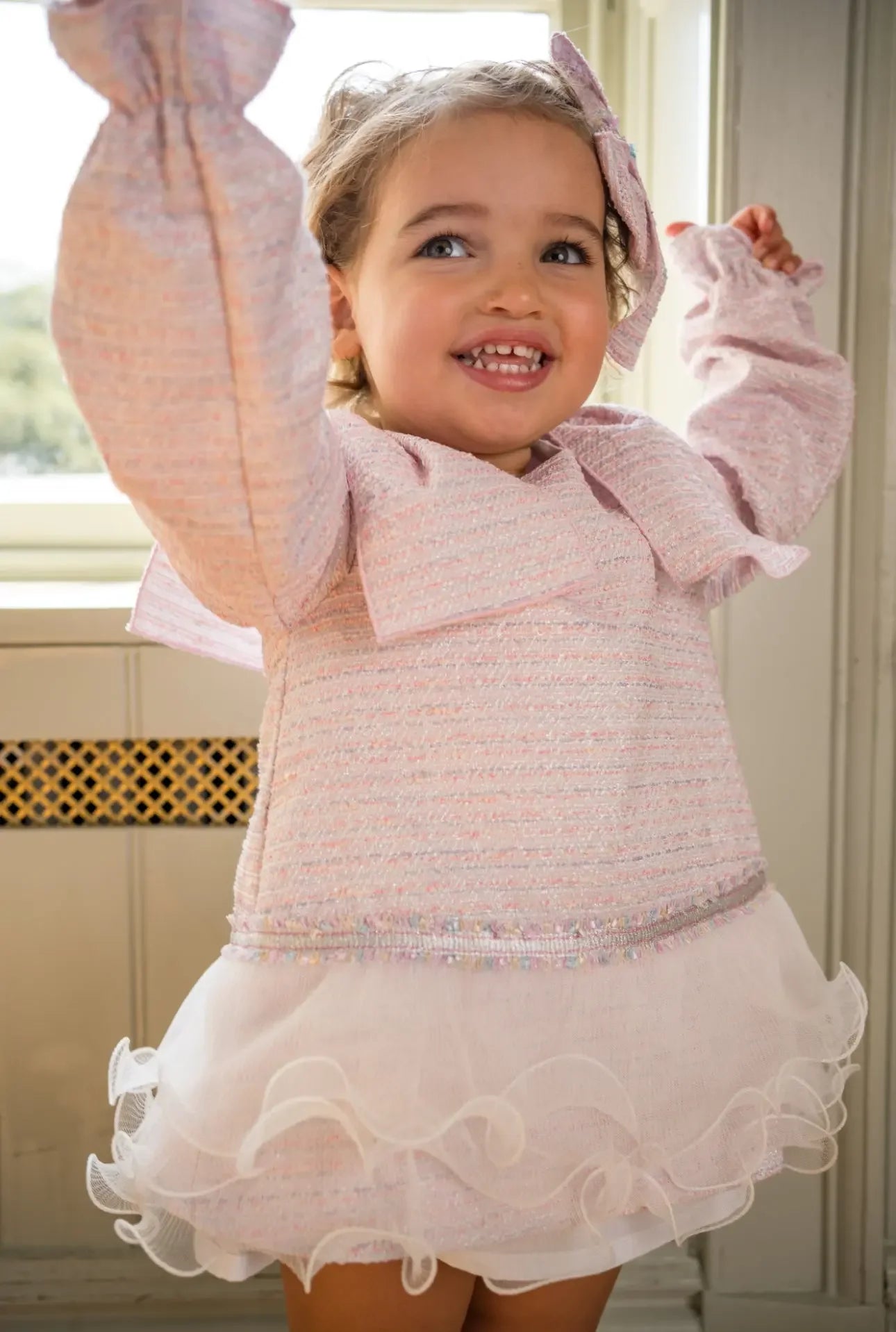 Smiling toddler girl in pink textured dress with ruffled tulle skirt and matching bow indoors