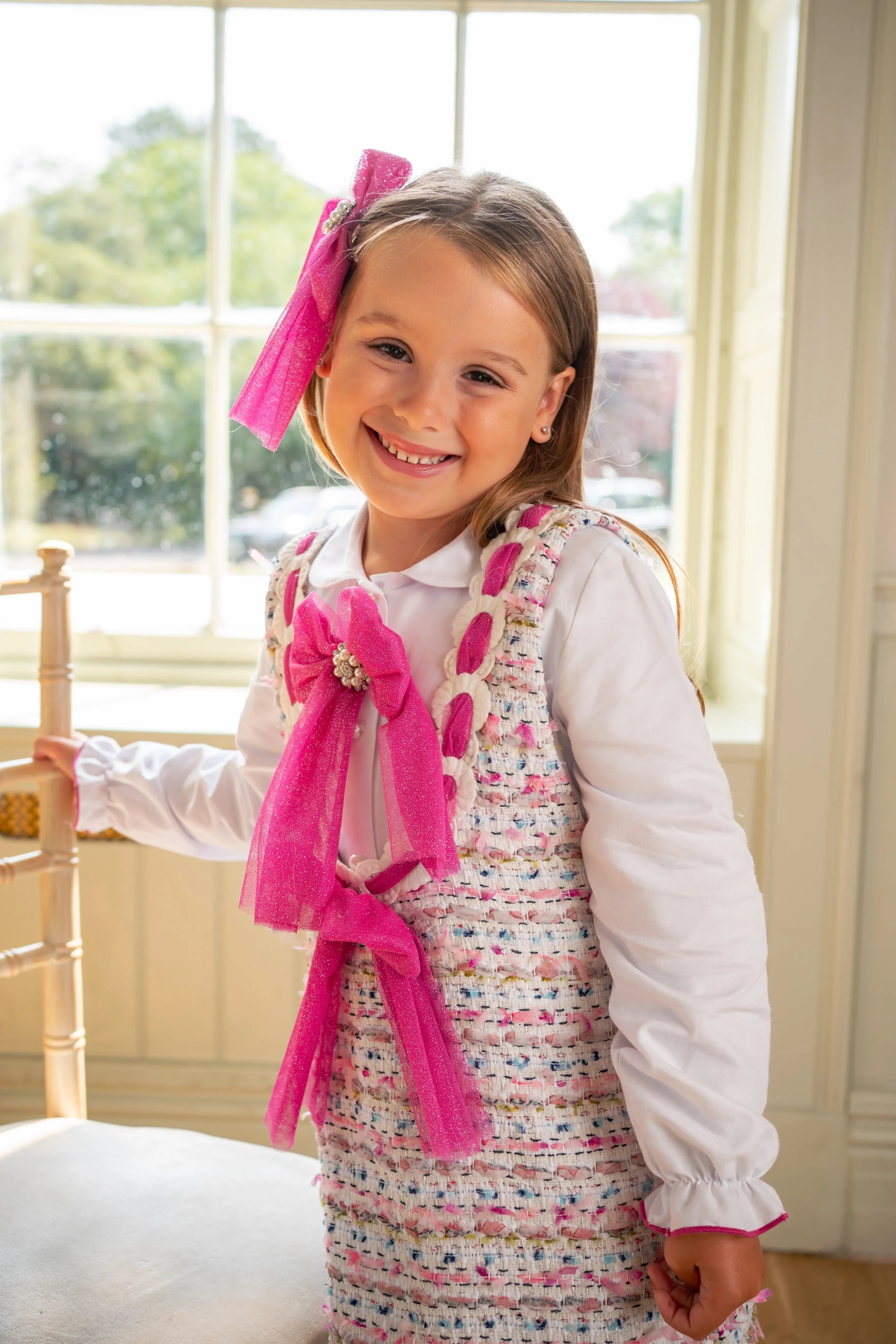 Smiling young girl wearing white blouse and textured dress with pink bows indoors by window