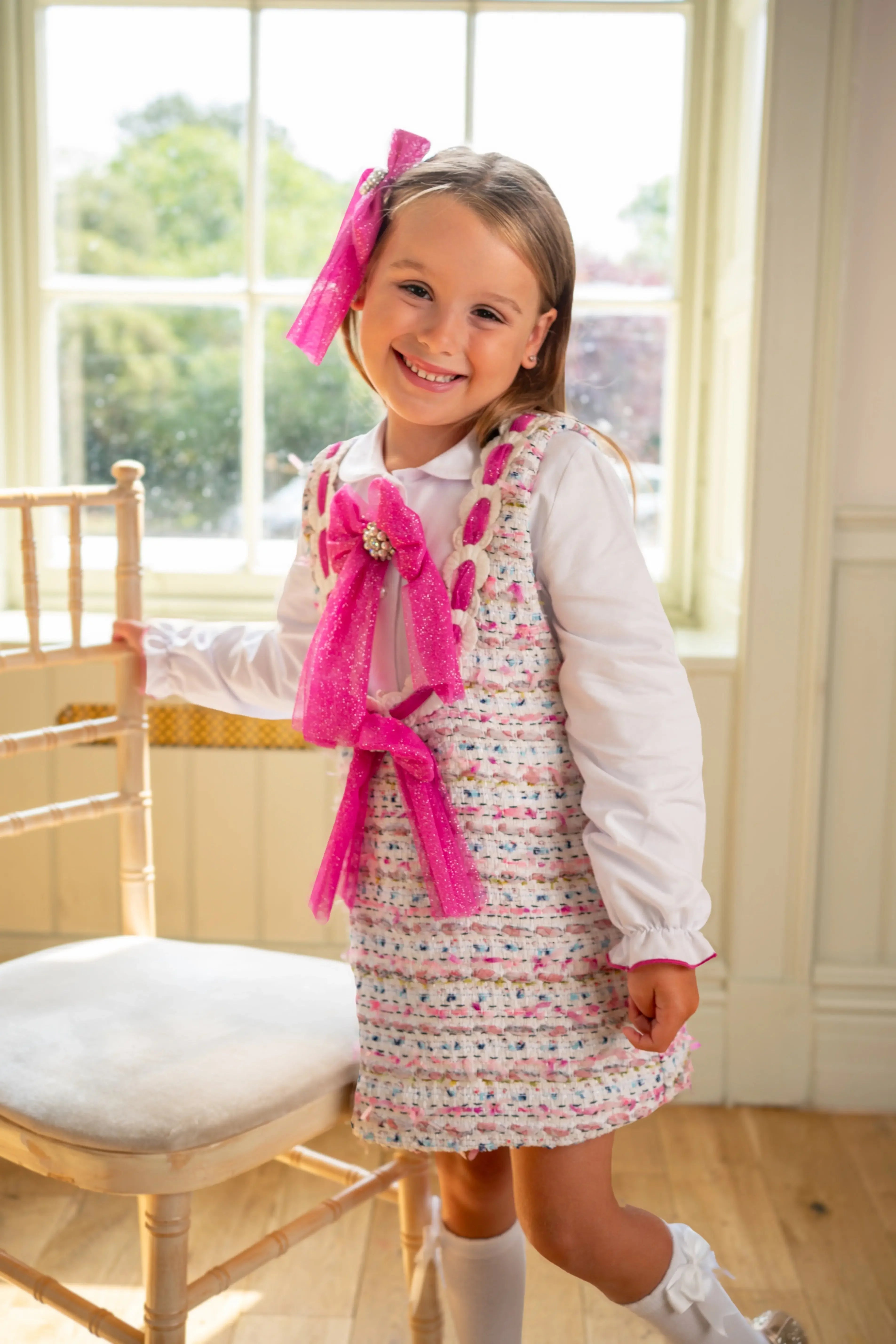 Smiling young girl in white blouse and pink bow tie dress standing by a wooden chair in a sunlit room