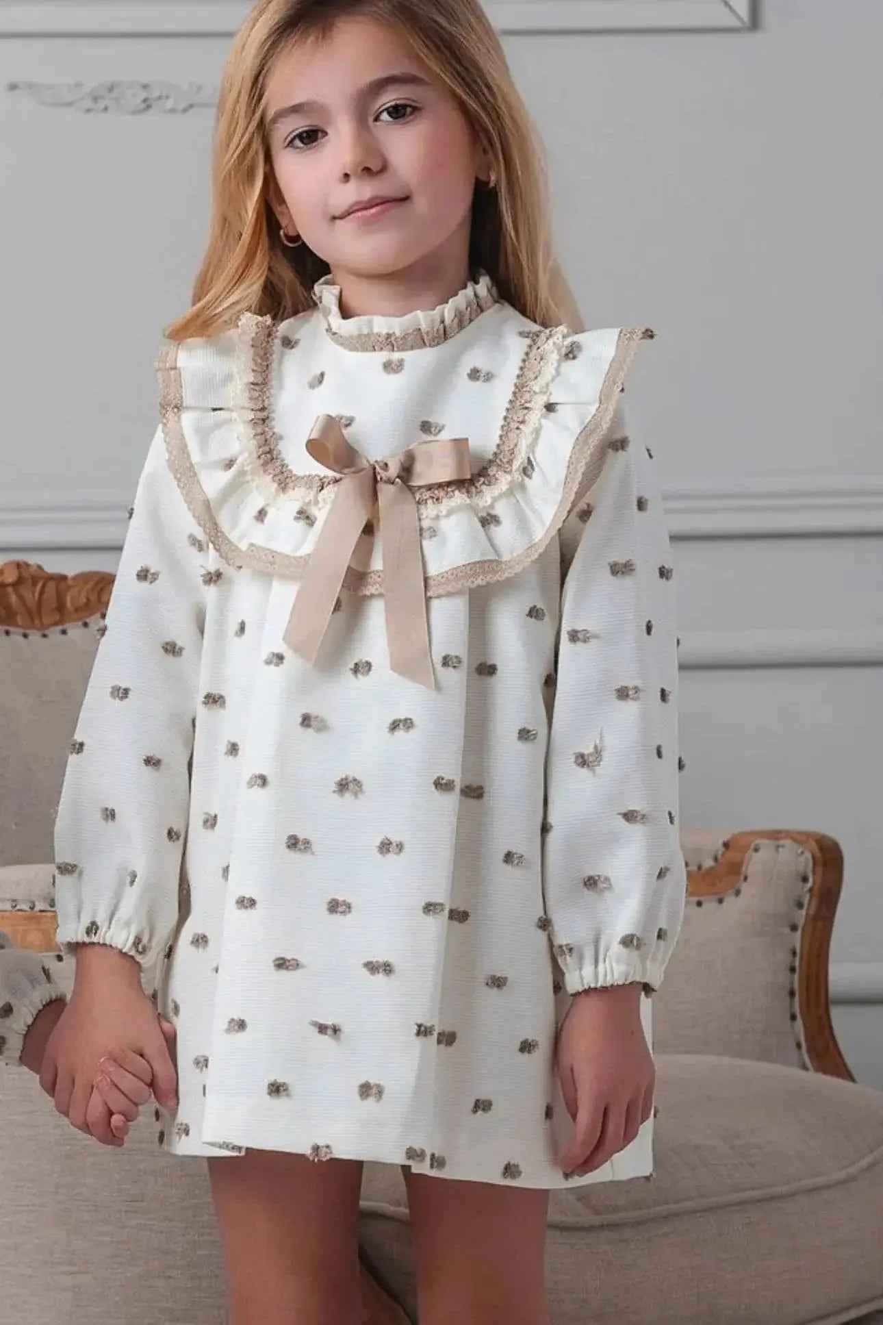 Young girl in a cream dress with camel spot pattern and ruffle collar bow, holding hands indoors