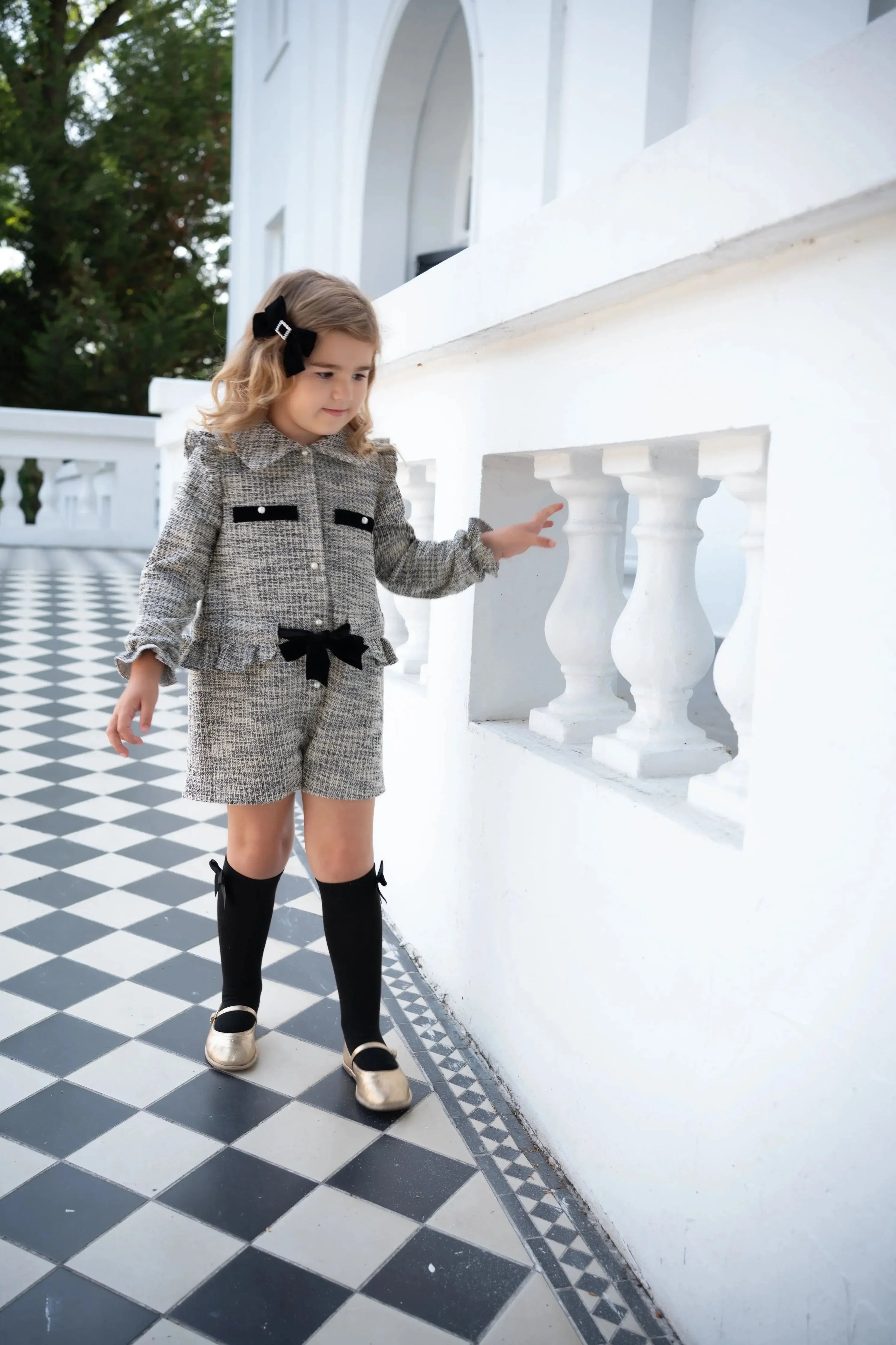 Little girl in black tweed jacket and shorts with black knee socks and gold shoes on checkered floor