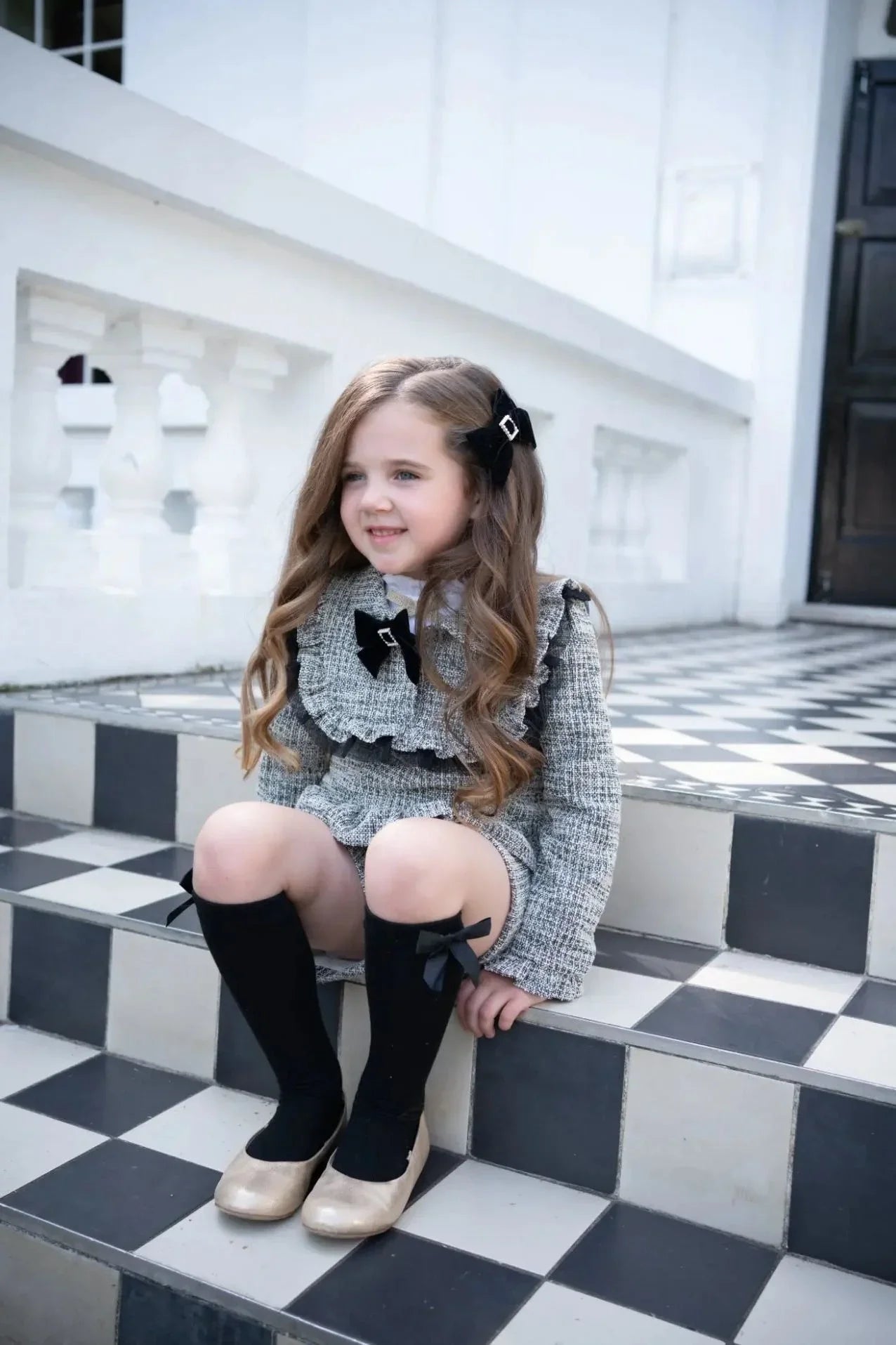 Little girl in black tweed short set with bows sitting on black and white checkered stairs