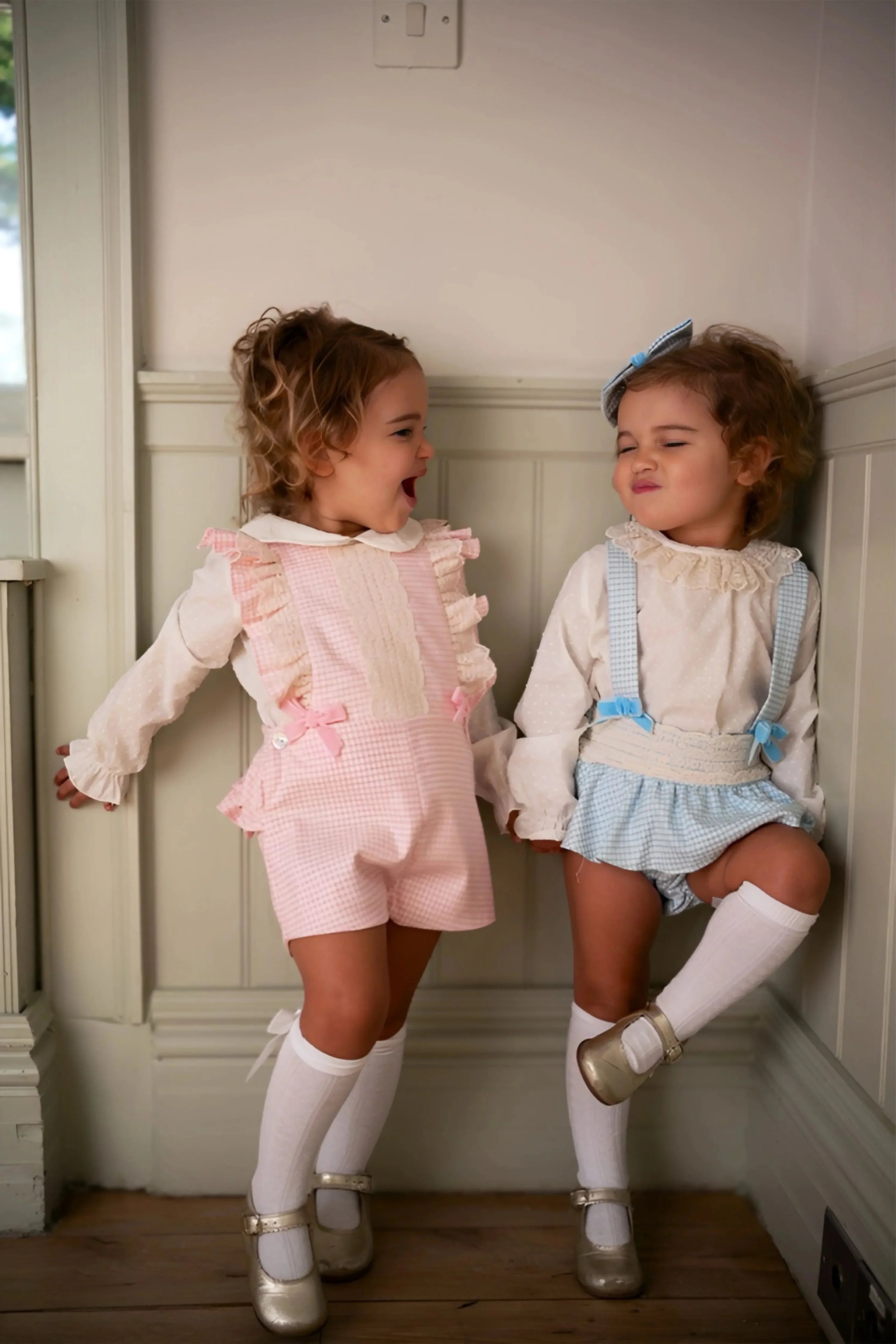 Two toddlers wearing pink and blue gingham romper sets with white blouses, knee-high socks, and gold shoes