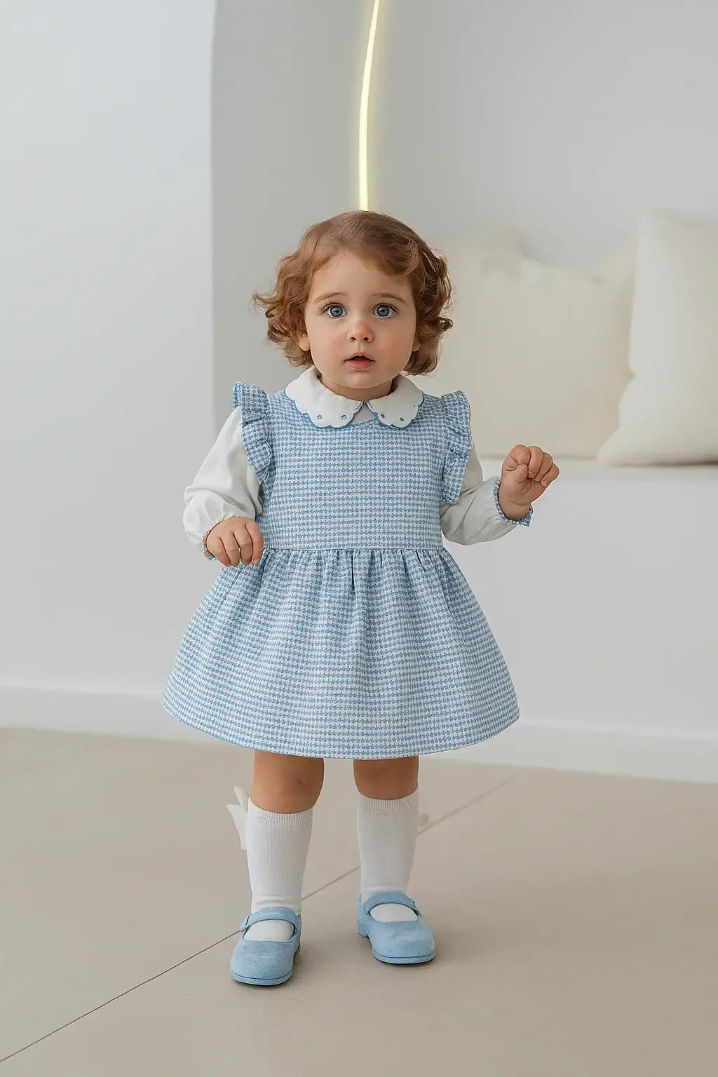 Toddler girl wearing blue pinafore dress with white blouse, white socks, and blue shoes in minimalist room