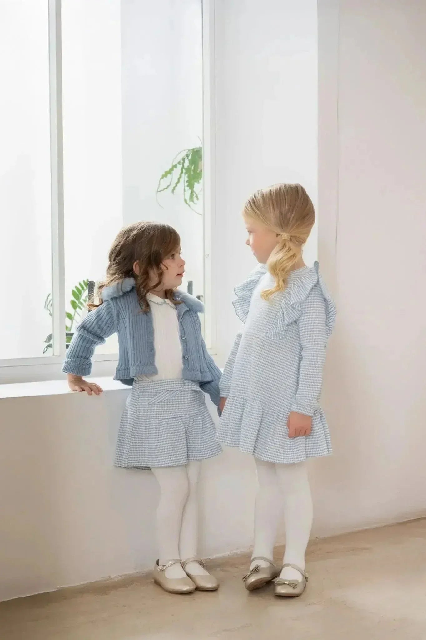 Two young girls wearing light blue checkered dresses and tights standing indoors by a window